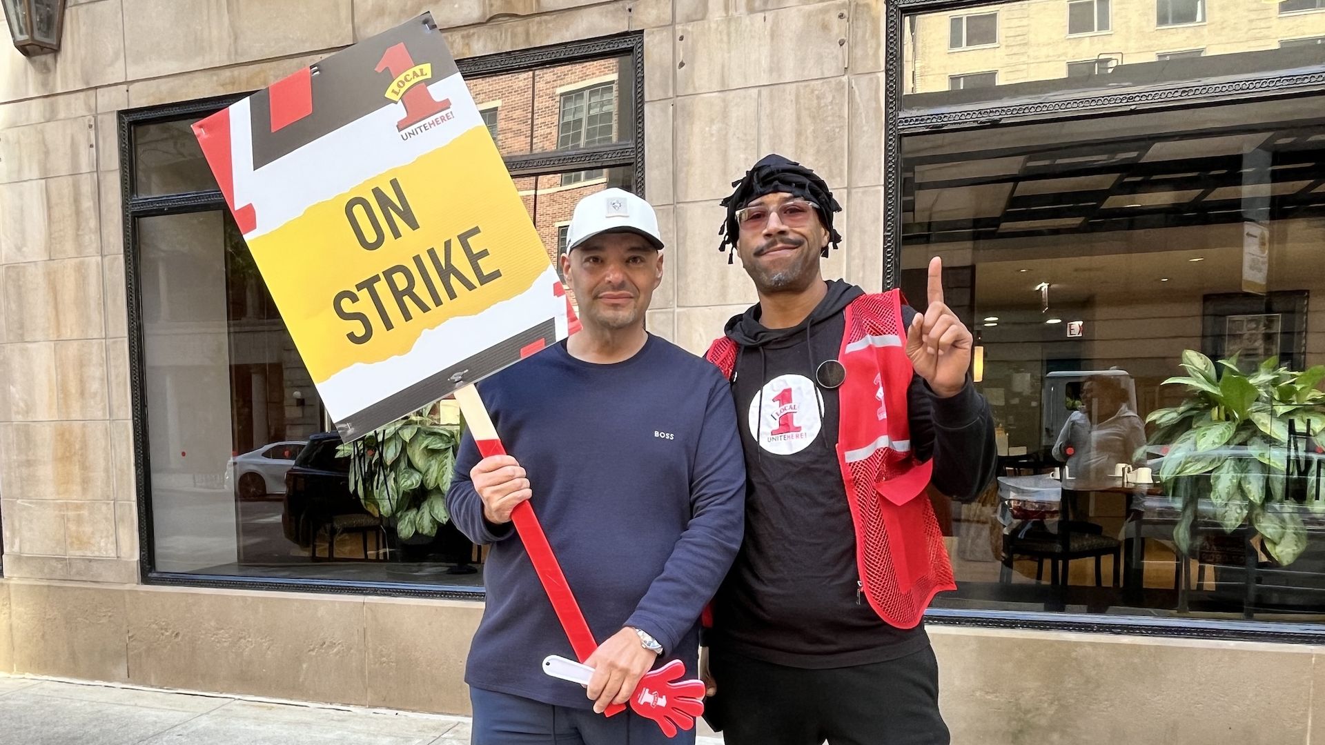 Two men stand outside a beige stone building; one holds a large sign reading "ON STRIKE" in bold yellow and black, while the other wears a red vest and matching pants, smiling and raising a finger.