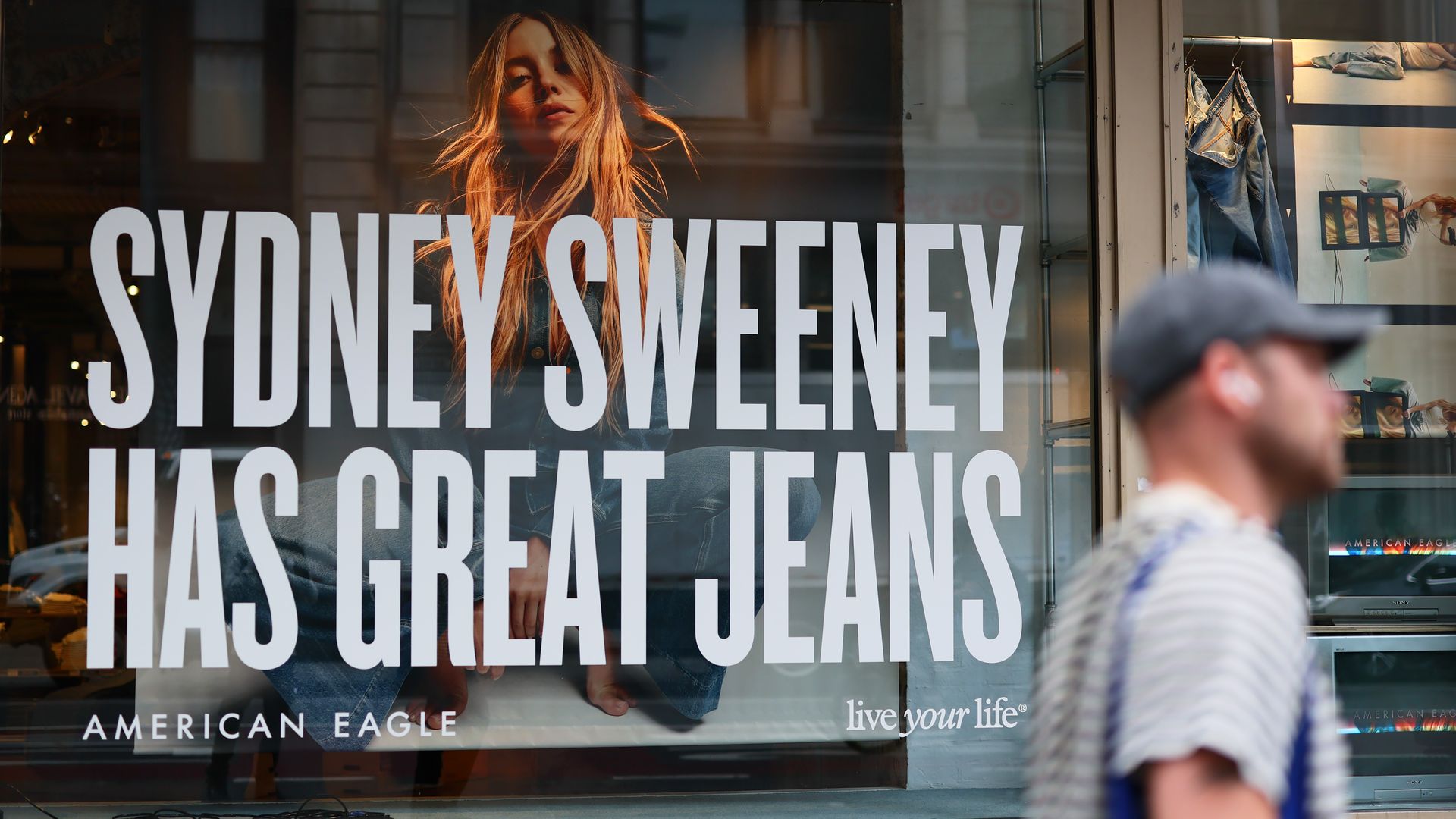 Store window displaying a large poster of a young woman in denim jeans with the text "SYDNEY SWEENEY HAS GREAT JEANS" from American Eagle, with a blurred man walking past.