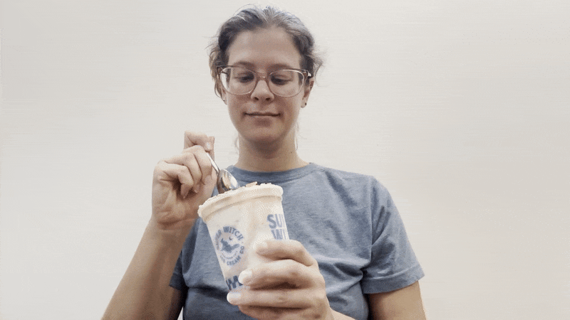 Person with glasses and gray shirt scooping ice cream from a white container with blue lettering against a plain light background.