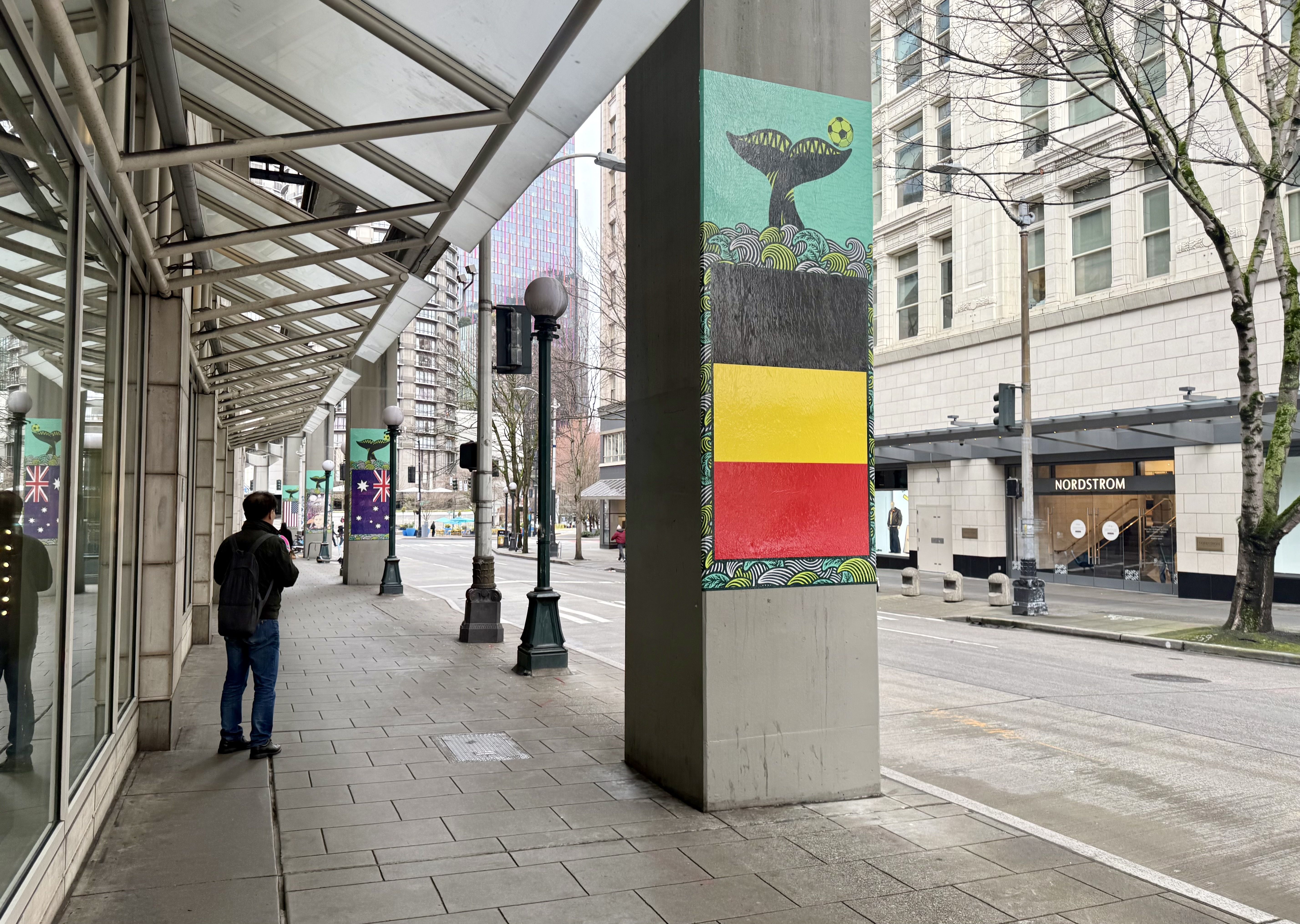 Downtown Seattle sidewalk with decorated pillars showing flag-themed art and a whale design, lined with buildings including a Nordstrom store, and a person standing near a glass window.