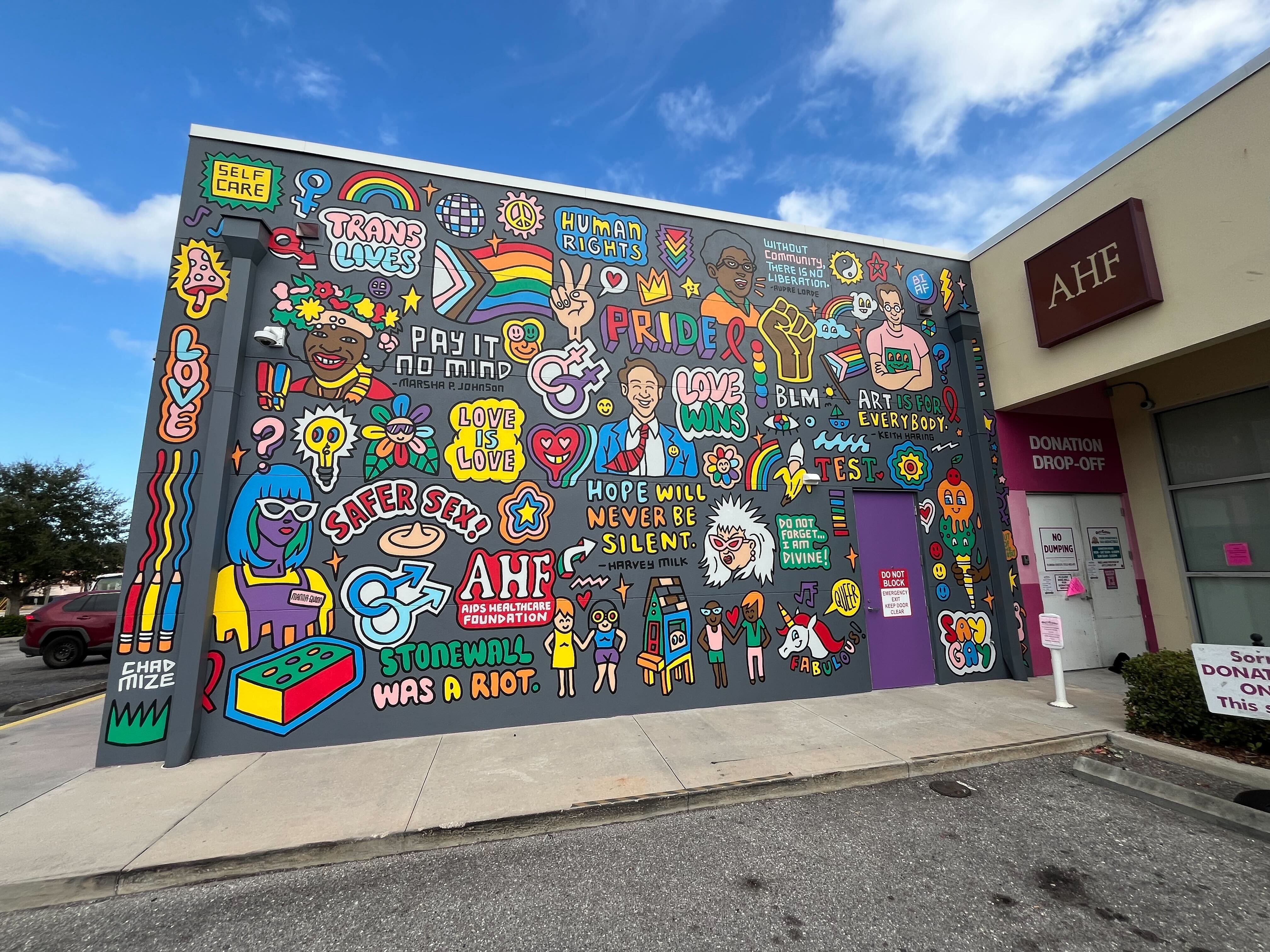 Colorful mural on AHF building featuring LGBTQ+ pride symbols, messages like "Love Wins," "Safer Sex!", "Stonewall Was a Riot," and portraits promoting self-care, human rights, and community.