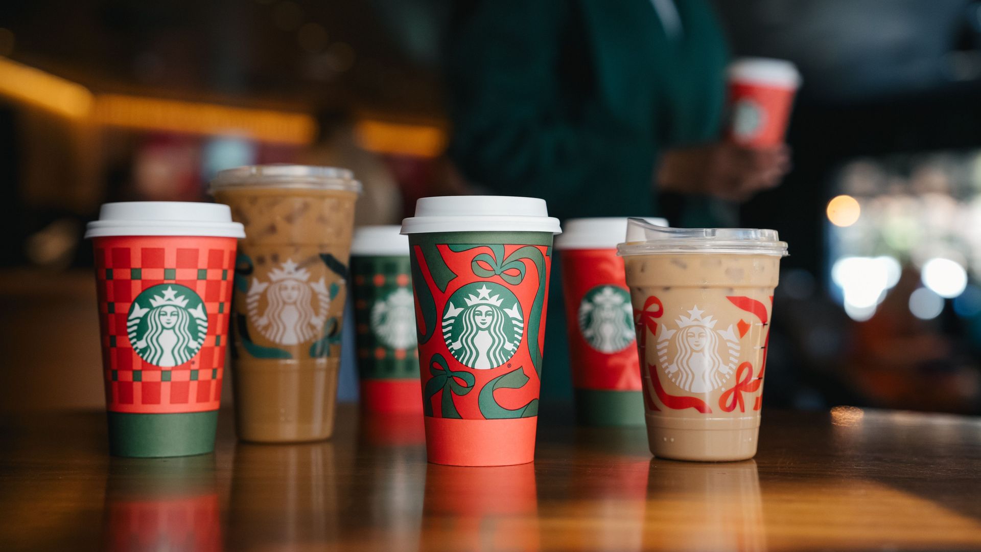 Six Starbucks holiday-themed cups on a wooden table, featuring red, green, and festive designs, some iced coffee; blurred background with person holding cup.