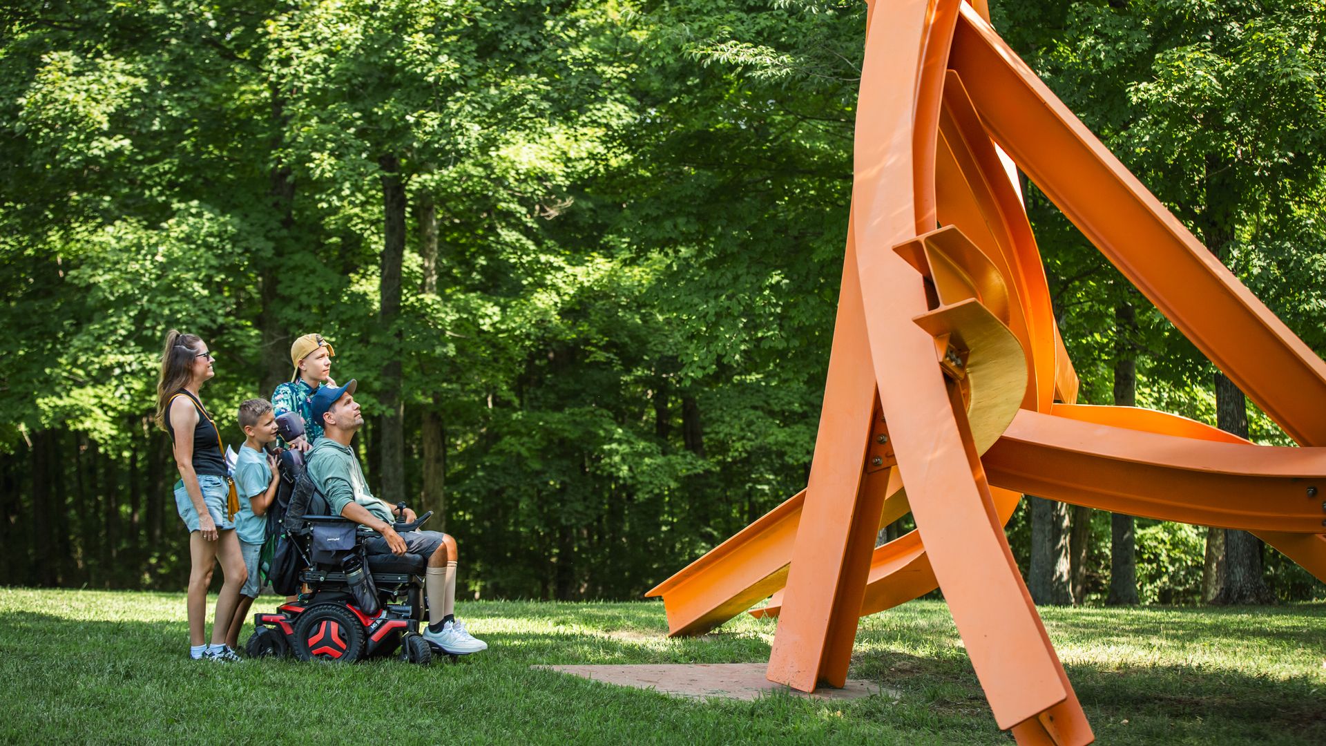 A family, including a man in a motorized wheelchair and several kids, watches a large orange geometric sculpture in a sunny, green park.