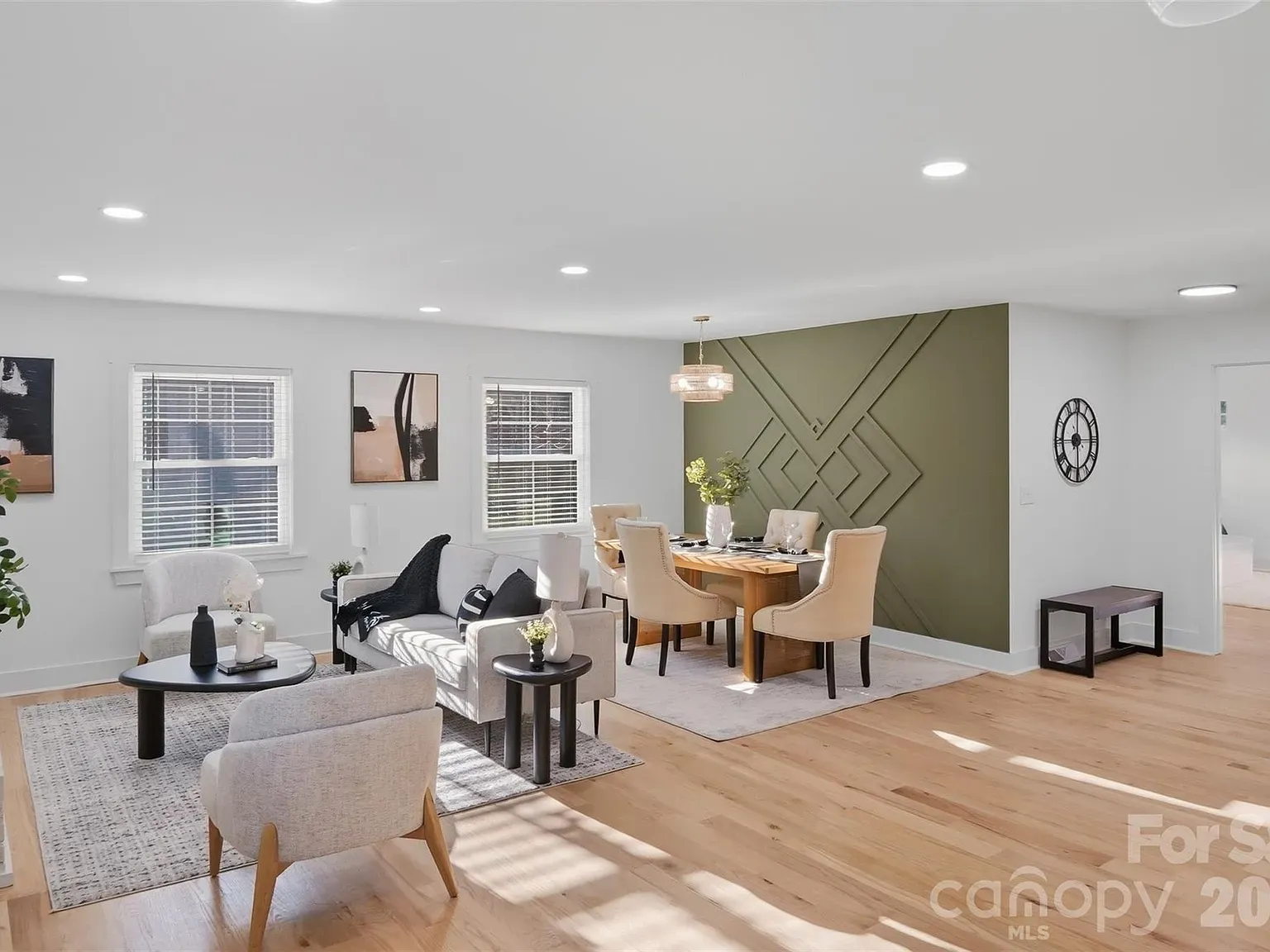 Bright open living and dining area with white walls, light hardwood floors, and recessed lights. A green accent wall with geometric panels behind a wooden dining table and beige chairs.