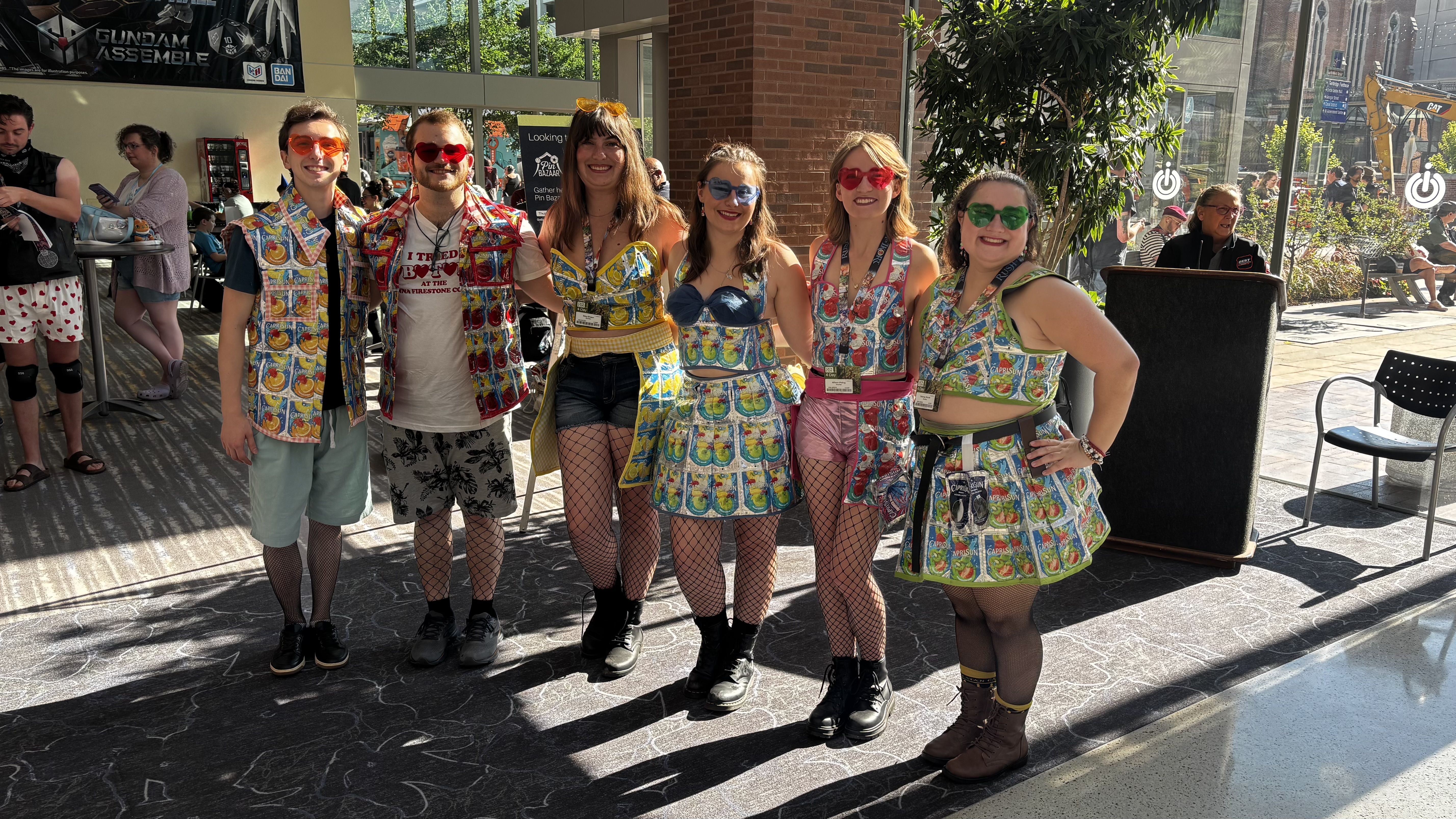 Group of six indoors, wearing bright Capri Sun pouch-pattern outfits and colorful heart-shaped sunglasses; they smile in a sunny atrium with banners and a construction site outside.