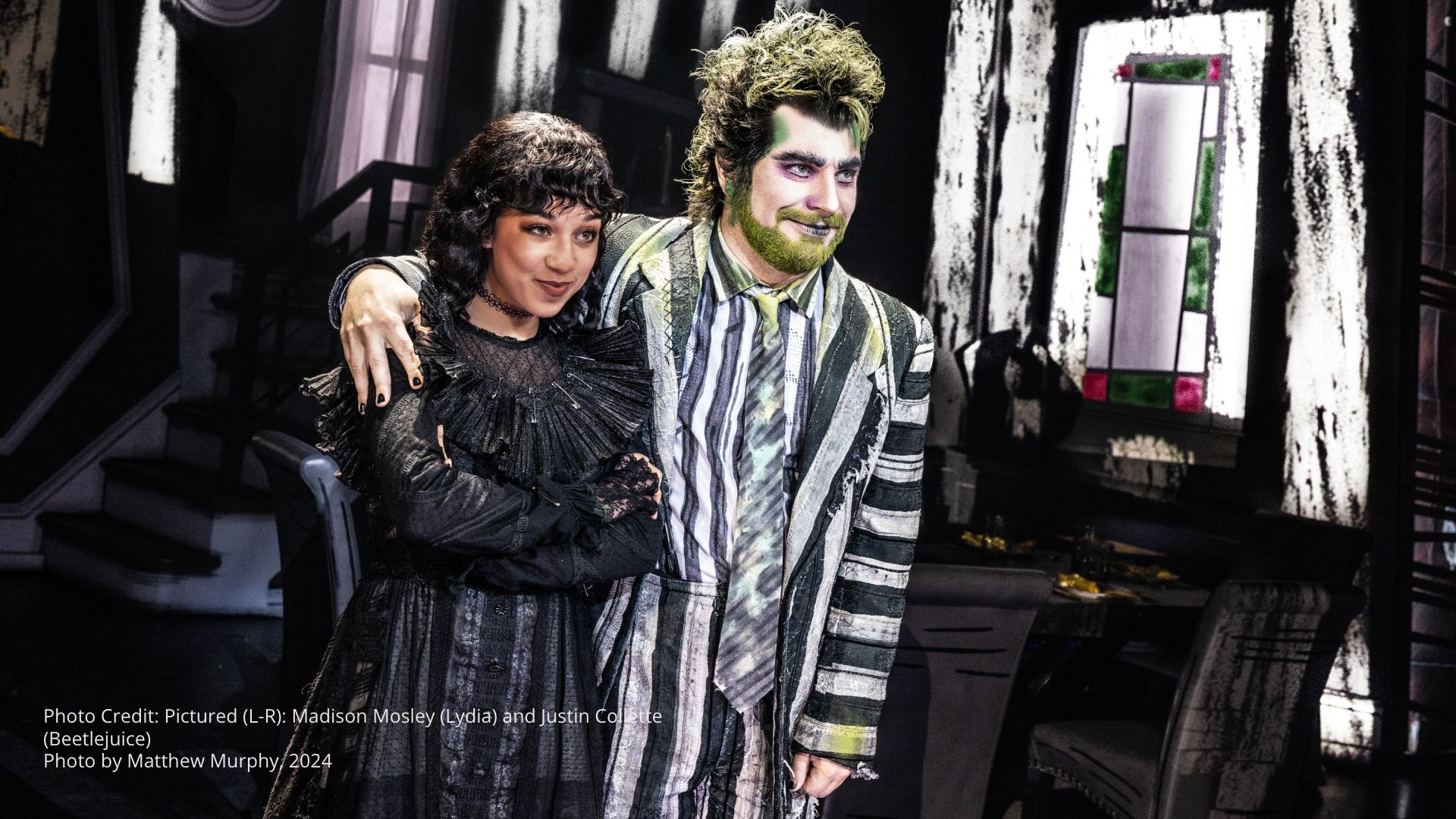 Two actors in black-and-white striped costume and dark gothic dress pose together on stage set with black-and-white patterns and stained glass window, portraying Beetlejuice and Lydia.