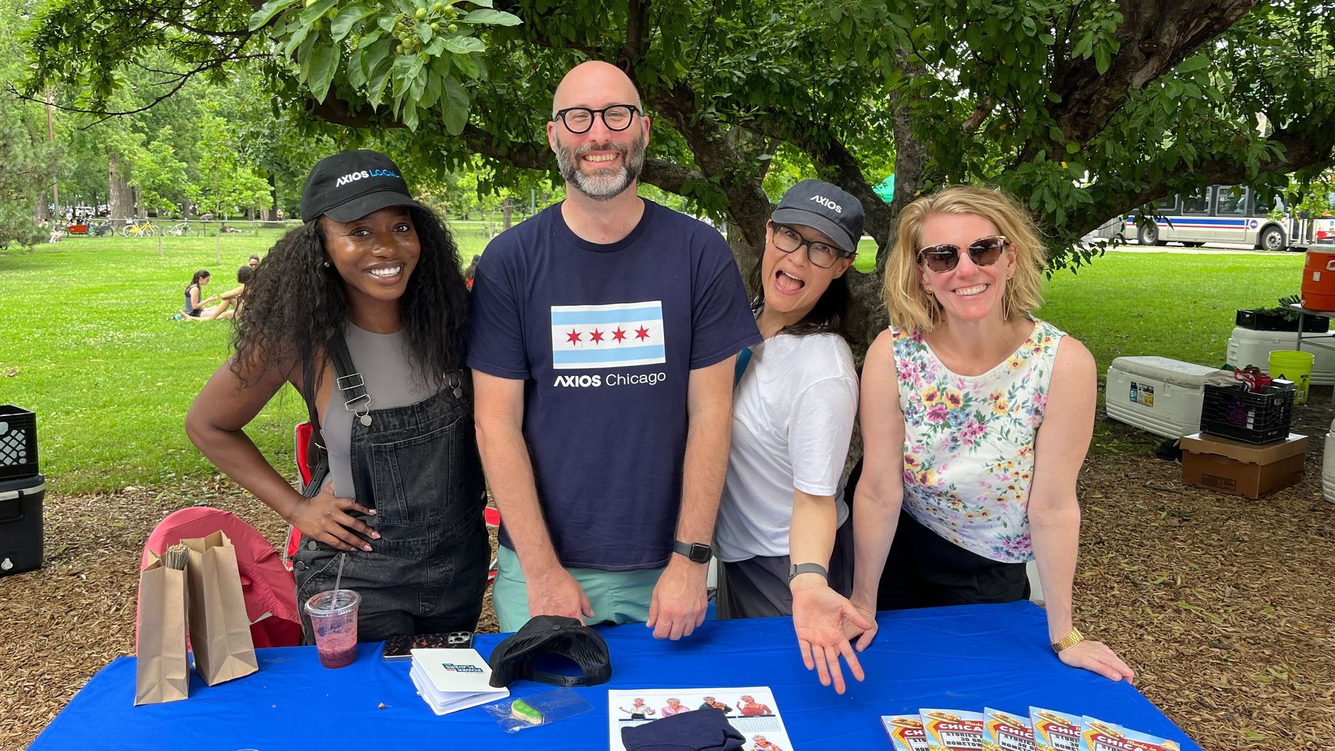 Four diverse people smiling behind a blue table with promotional materials, under a green leafy tree in a park setting, two wearing Axios hats and one an Axios Chicago shirt.