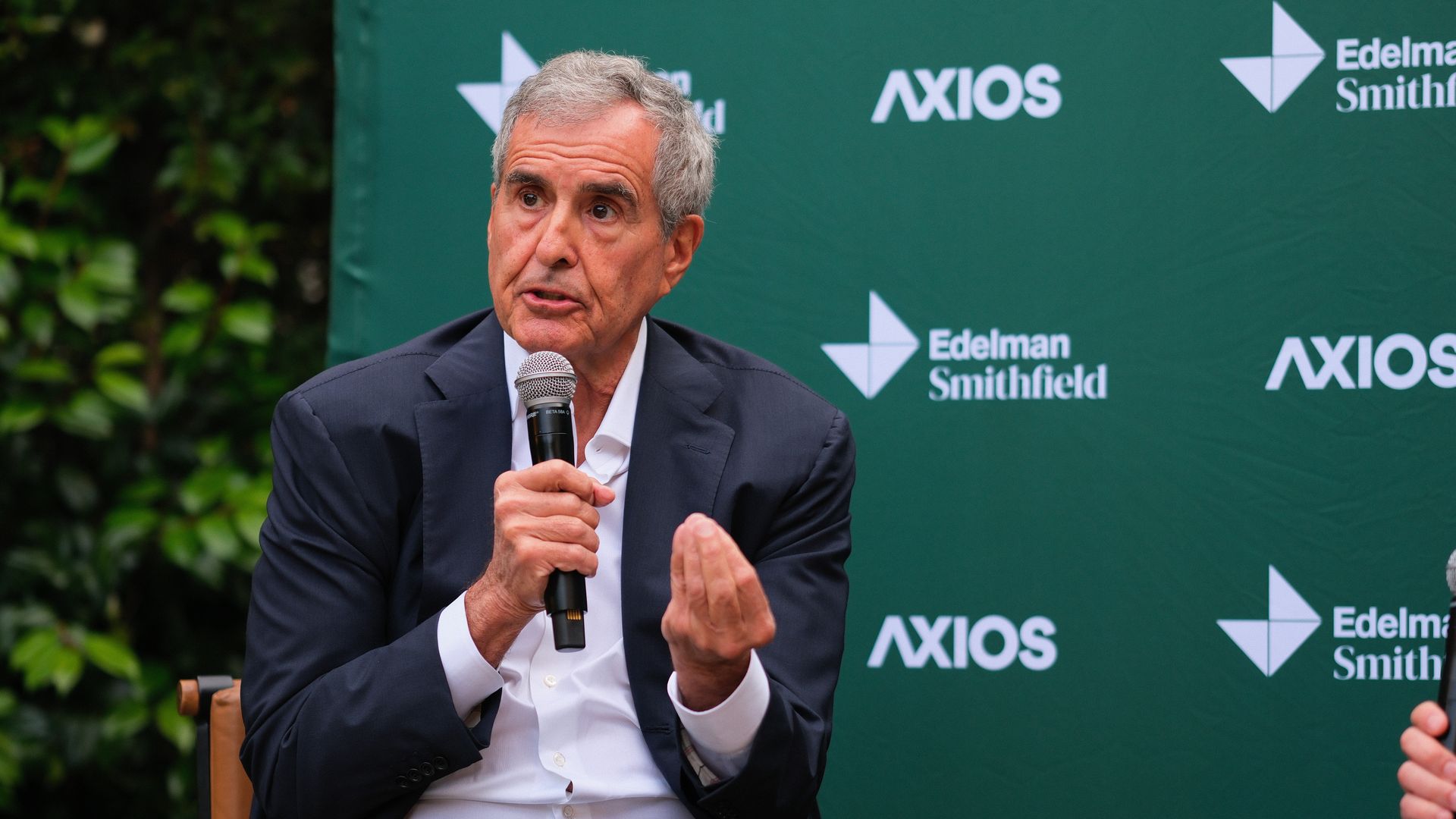 An older man with gray hair wearing a dark suit and white shirt speaks into a handheld microphone during a panel, seated before a green backdrop with the AXIOS and Edelman Smithfield logos.