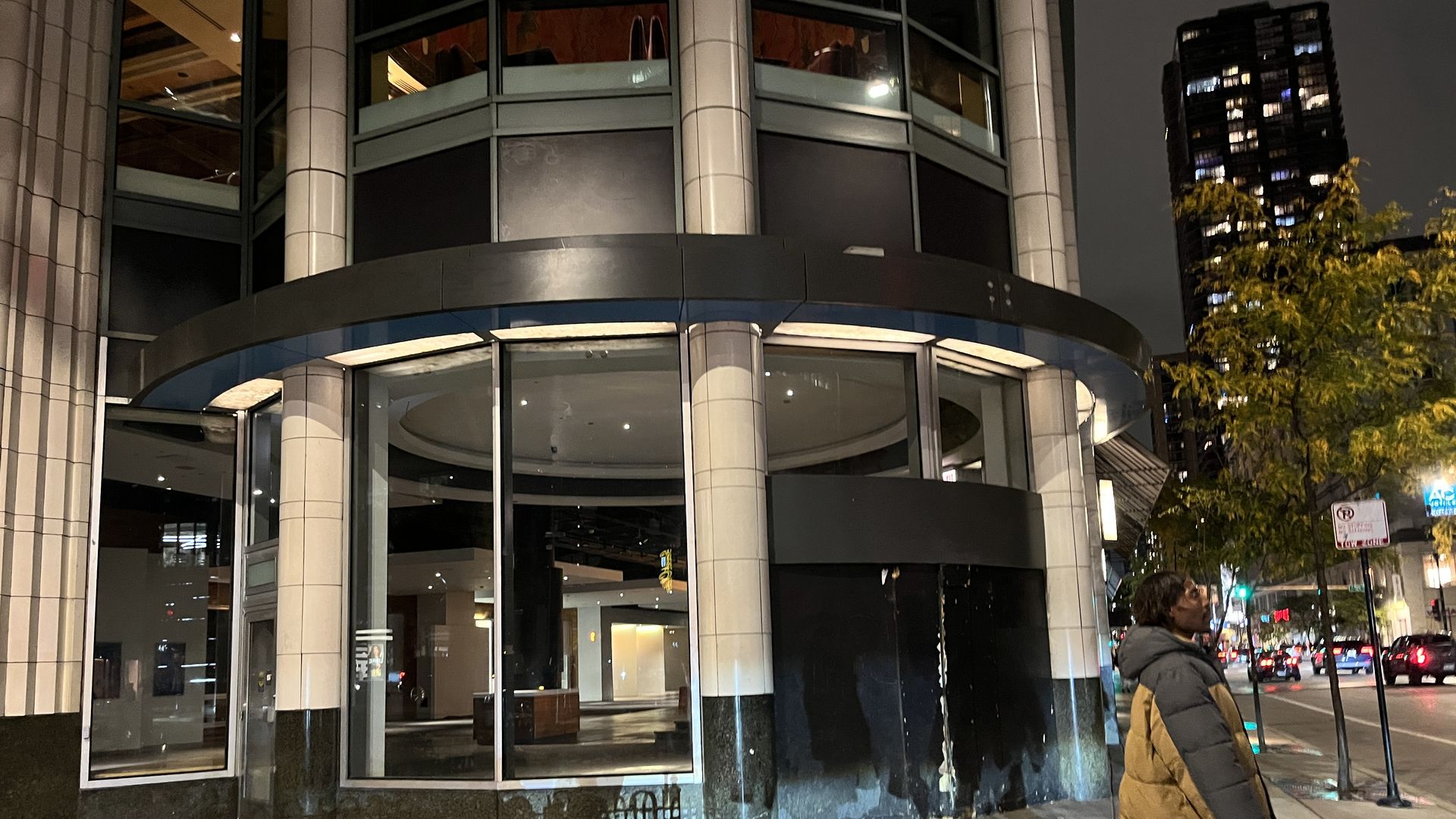 Nighttime city street scene with a person in a black and tan puffer jacket standing near a large glass building facade. Trees, parked cars, and lit windows are visible in the background.