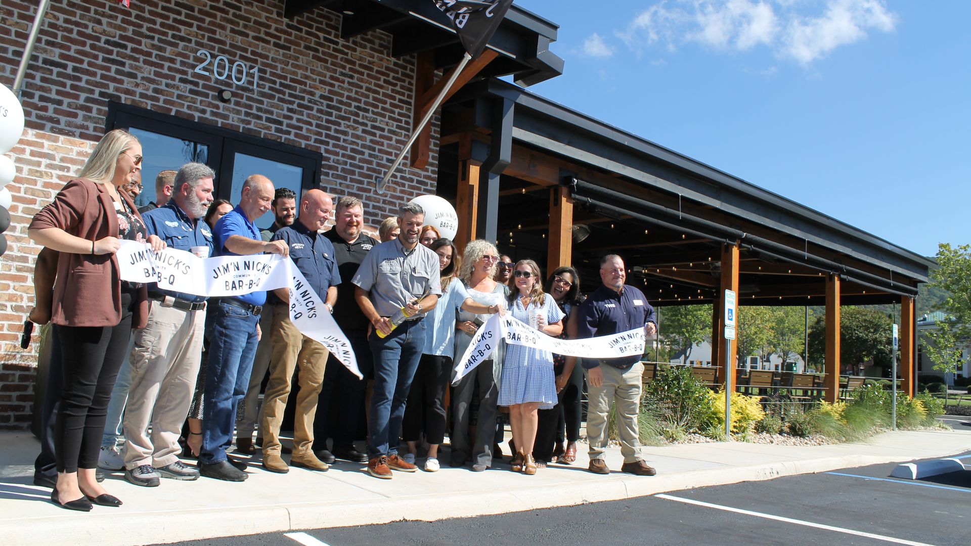 Group of people standing in front of a brick building with a sign reading "2001" holding a ribbon for Jim'N Nick's Bar-B-Q under a clear blue sky, celebrating a ribbon-cutting event.