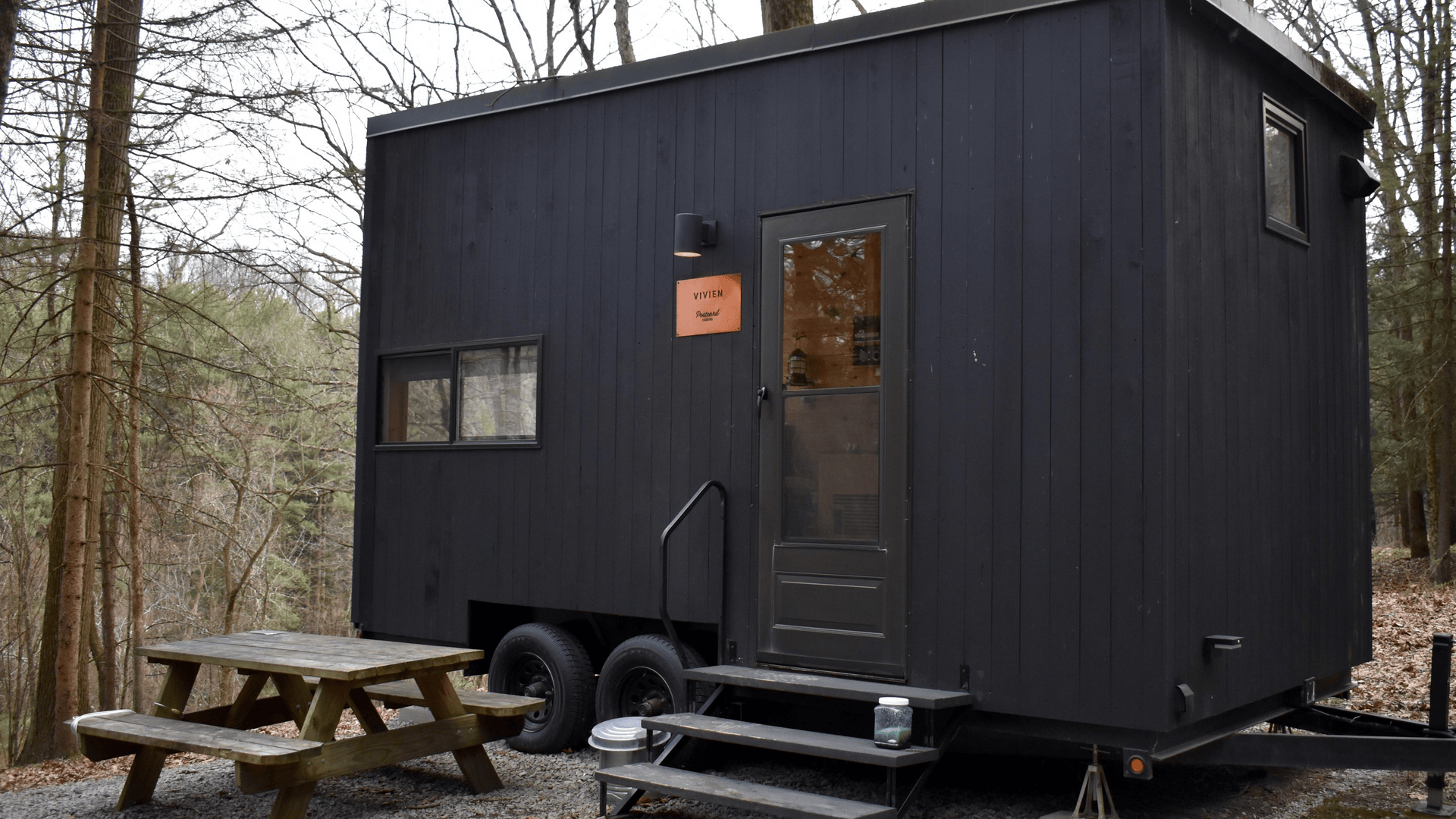 Dark tiny house on a trailer in a forest clearing. Front door with window, small steps, railing, and a wooden picnic table in the gravel foreground; leafless trees surround.