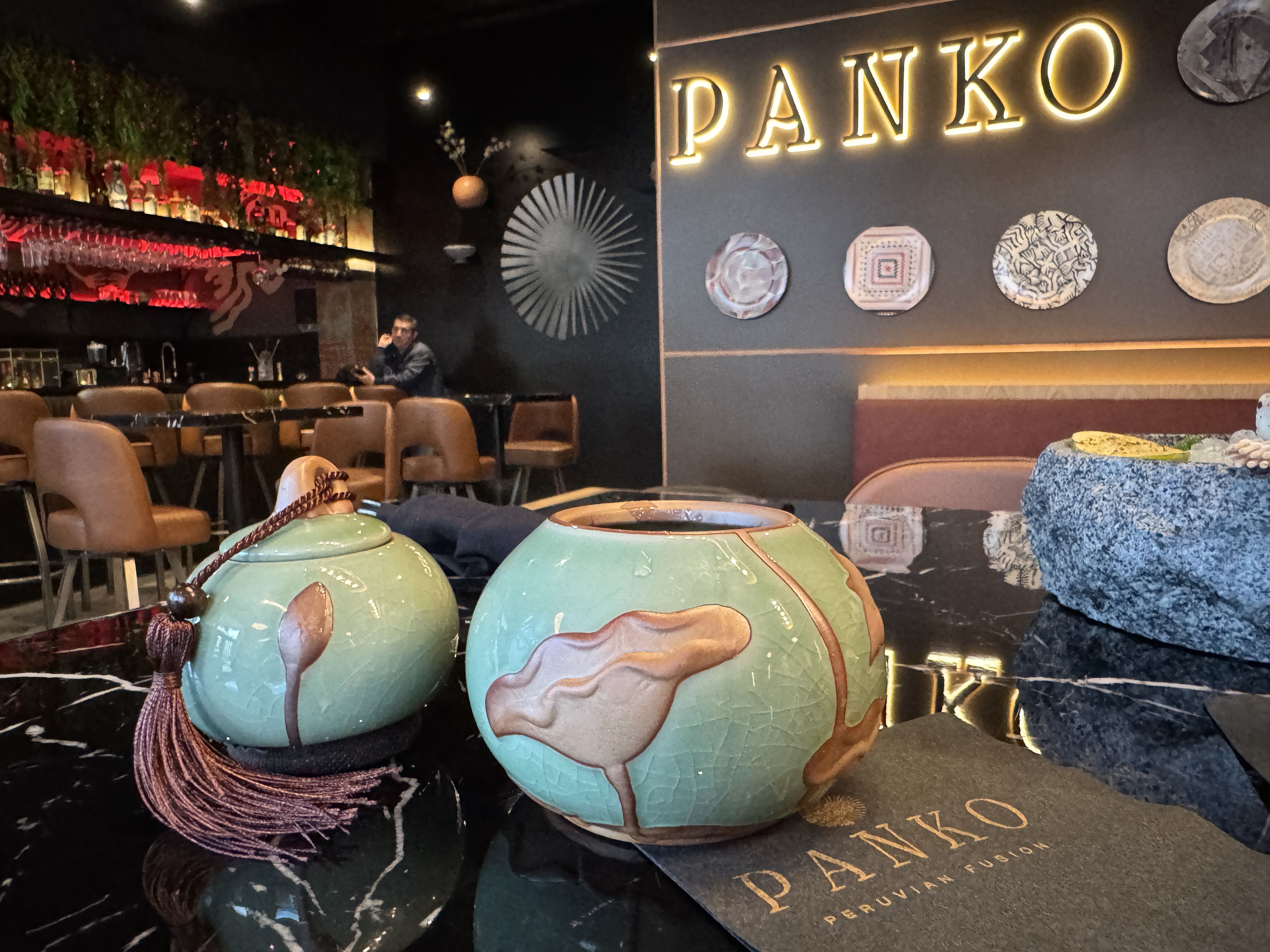 Interior of PANKO Peruvian Fusion restaurant with green ceramic pots on black marble table, brown chairs, red-lit bar, and decorative plates on dark wall.