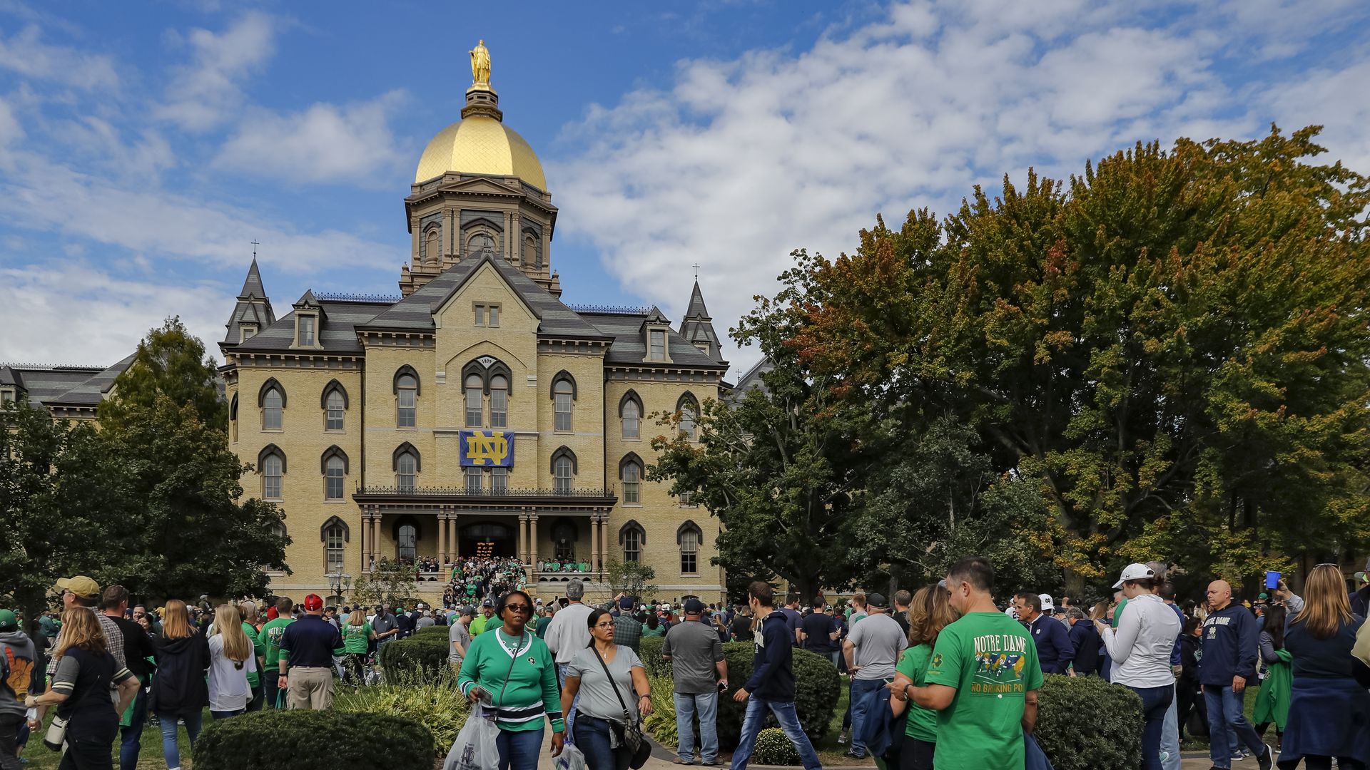 Crowd of people in green shirts on a campus plaza in front of a beige brick building with a gold dome and Notre Dame banner; blue sky and trees behind.