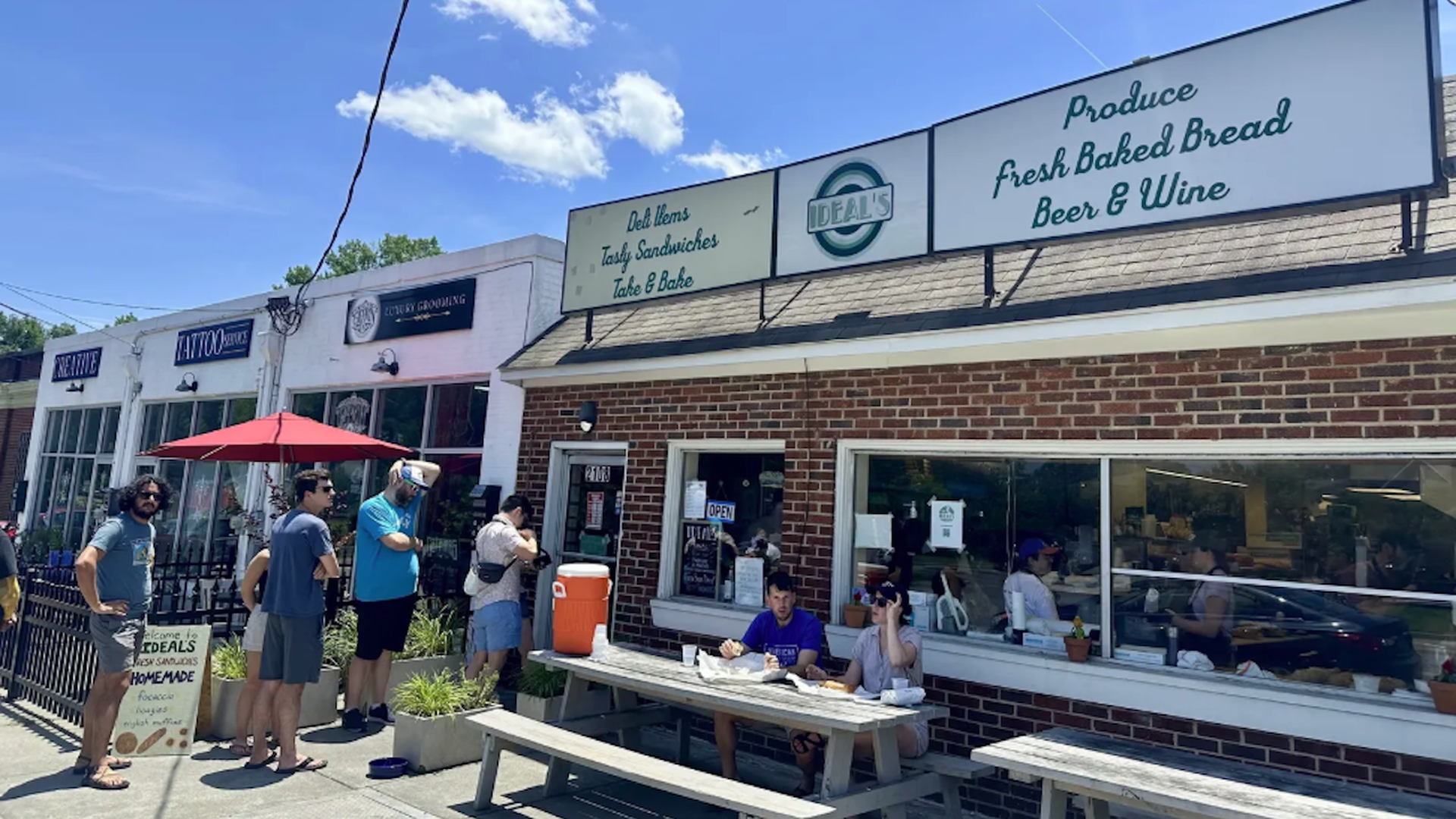 People standing in line outside a brick sandwich shop named Ideal's with signs for deli items, sandwiches, fresh baked bread, beer & wine on a sunny day with a blue sky.