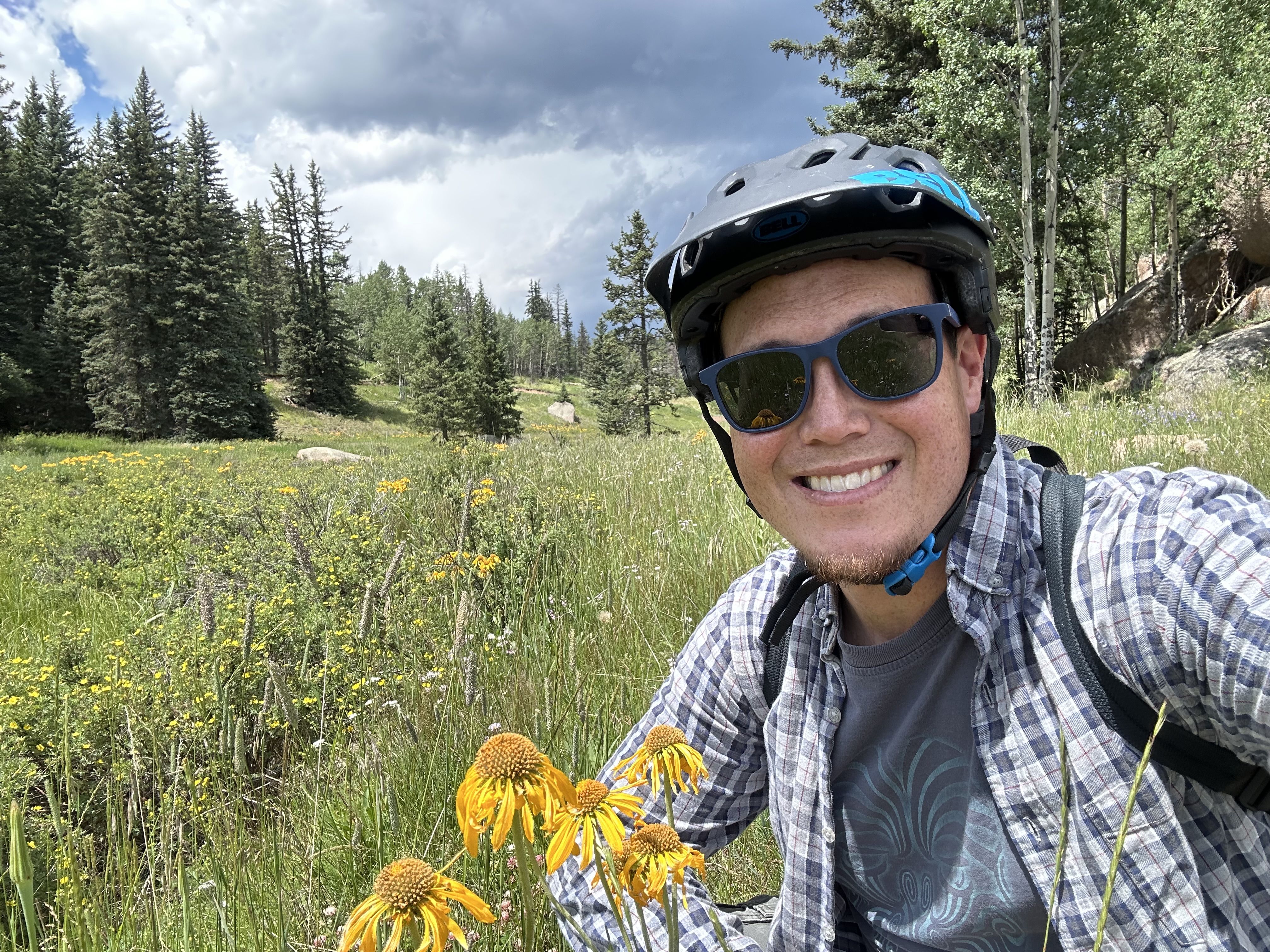 Man wearing a black bicycle helmet, blue sunglasses, and plaid shirt smiles while sitting in a green meadow with yellow flowers under a cloudy sky surrounded by pine trees.