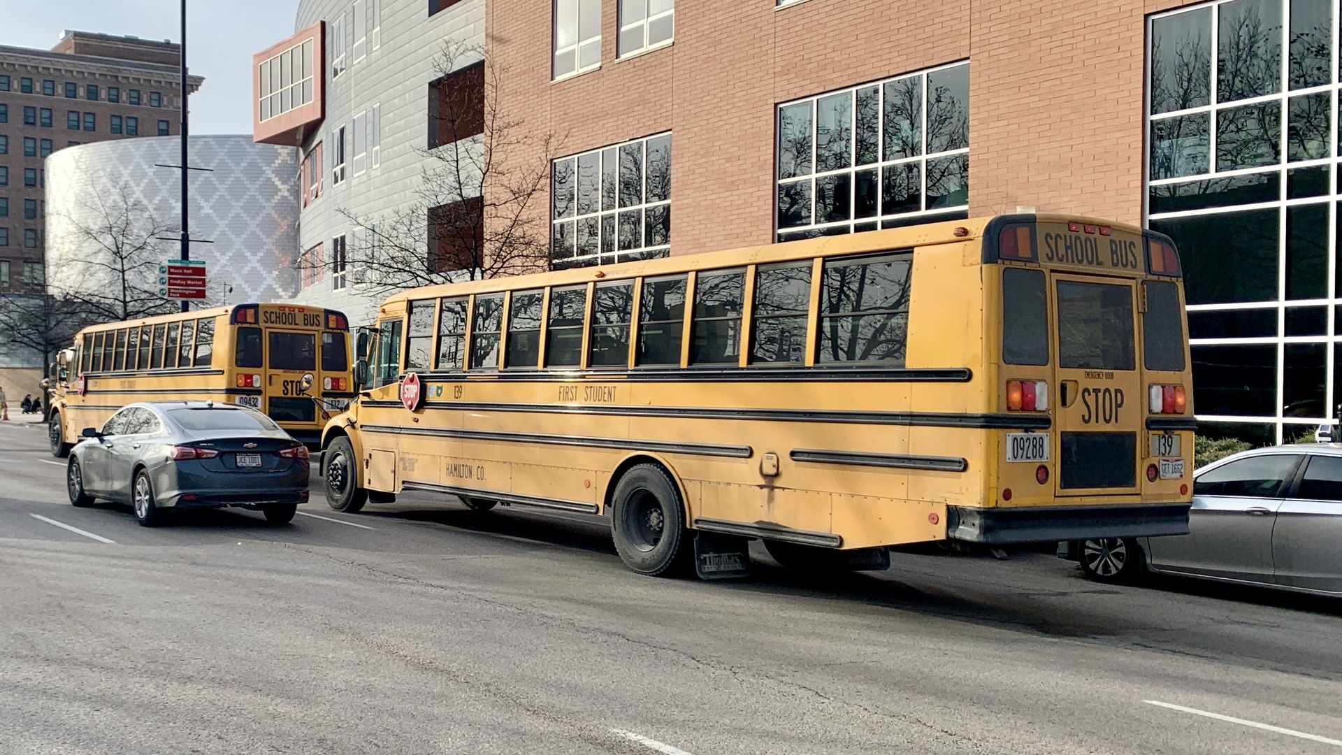 Street scene with two yellow school buses beside a modern brick and glass building; gray curved facade, leafless trees, a tall lamppost, and a silver car in the foreground.