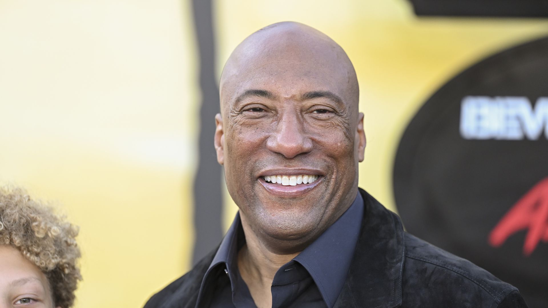 Smiling bald man wearing a black shirt and jacket stands in front of a blurred yellow and black background with red and white text.