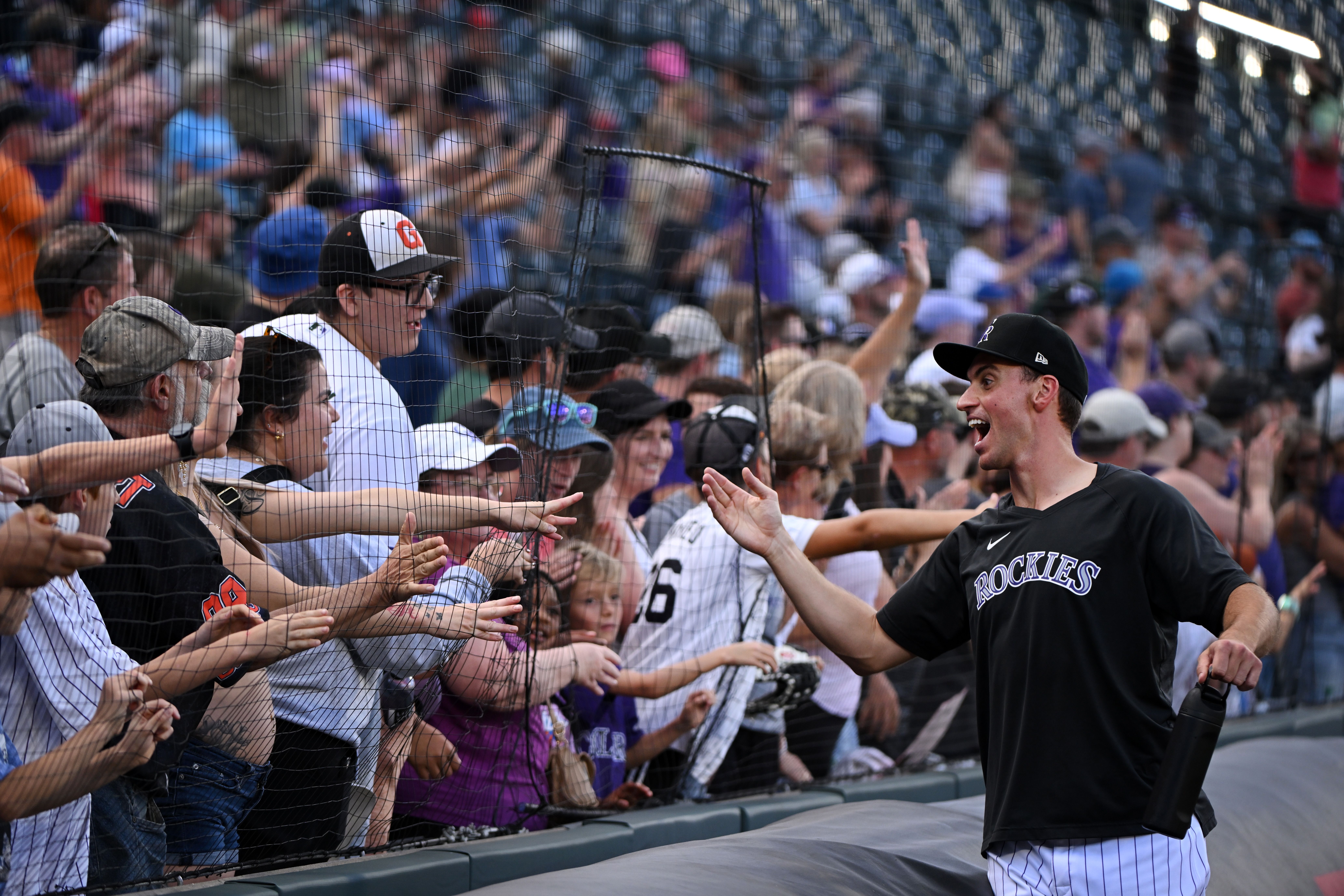 A man in a black shirt with the words ROCKIES on it attempts to touch hands with multiple people behind a netting inside a baseball stadium.