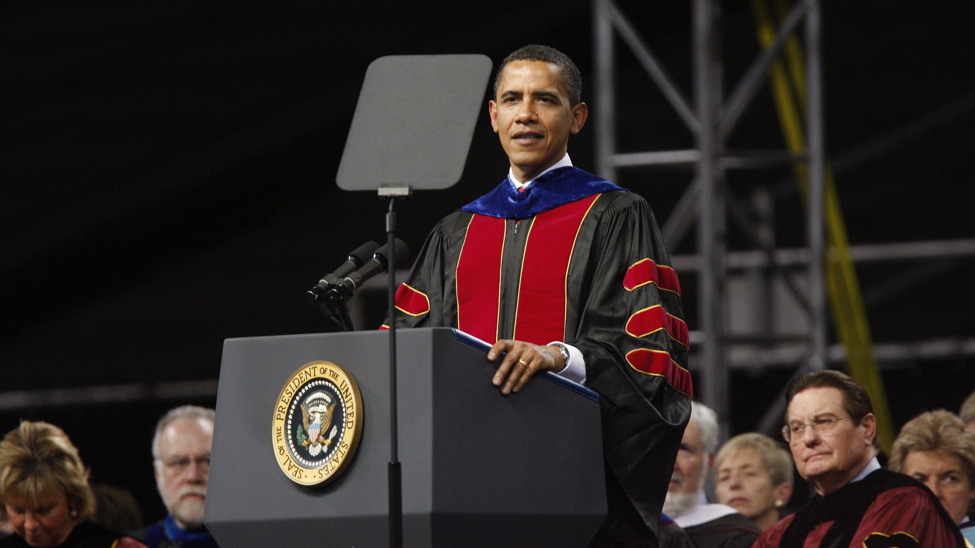 President Barack Obama wears academic robes while speaking at a lectern with the presidential seal on the front. Other people in academic robes are seated behind him.