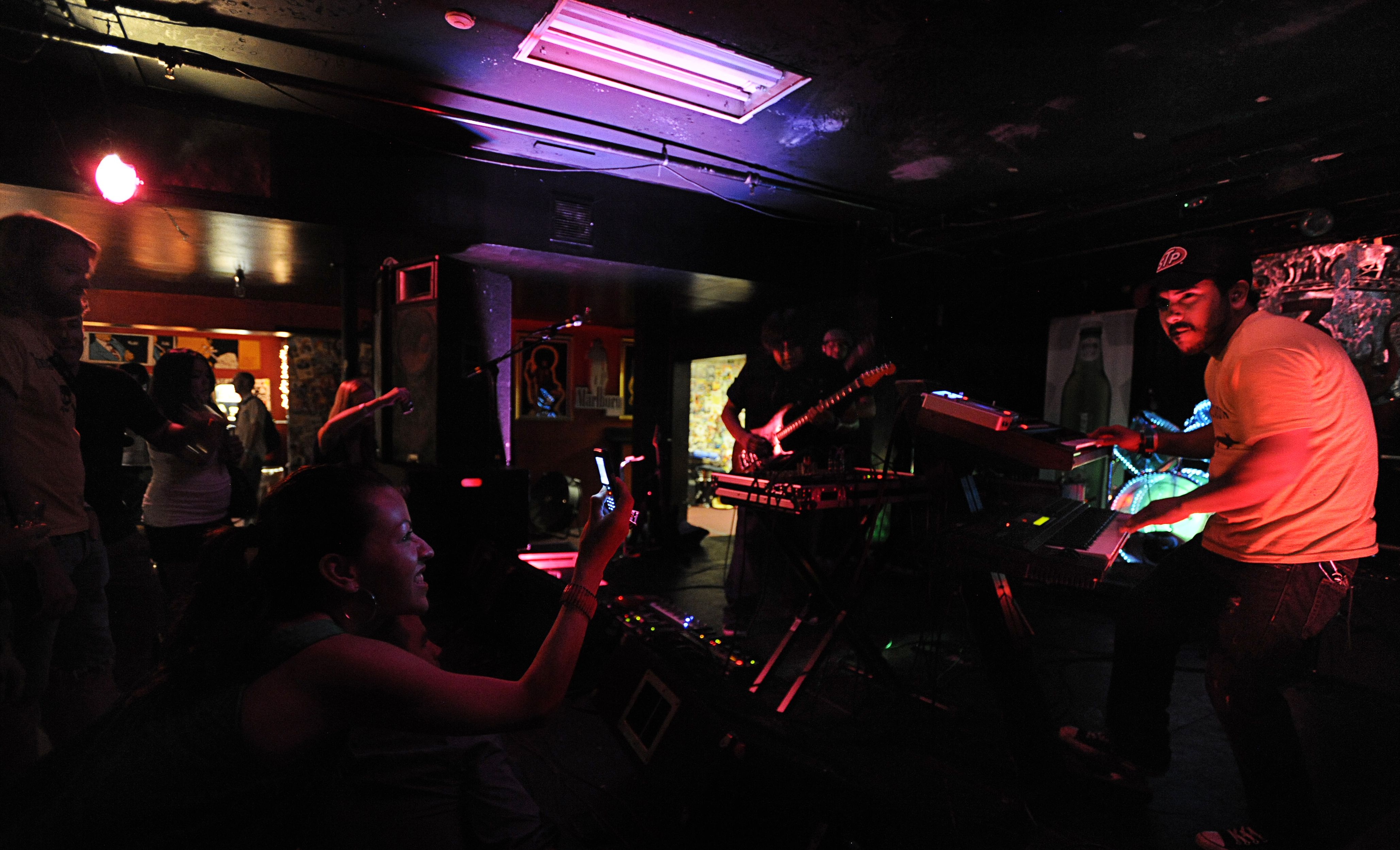 Dimly lit bar stage with a guitarist and keyboardist under pink and purple lights; a small crowd watches from the front, walls covered with posters and neon signs.