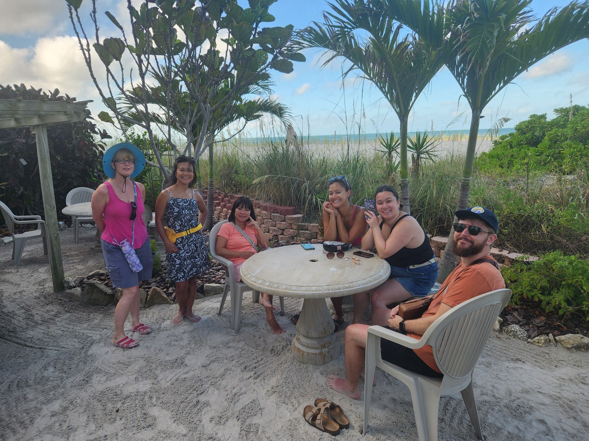 A group of people sitting at a table and smiling at the camera. In the background are lush plants, a sandy beach and a thin strip of blue water.