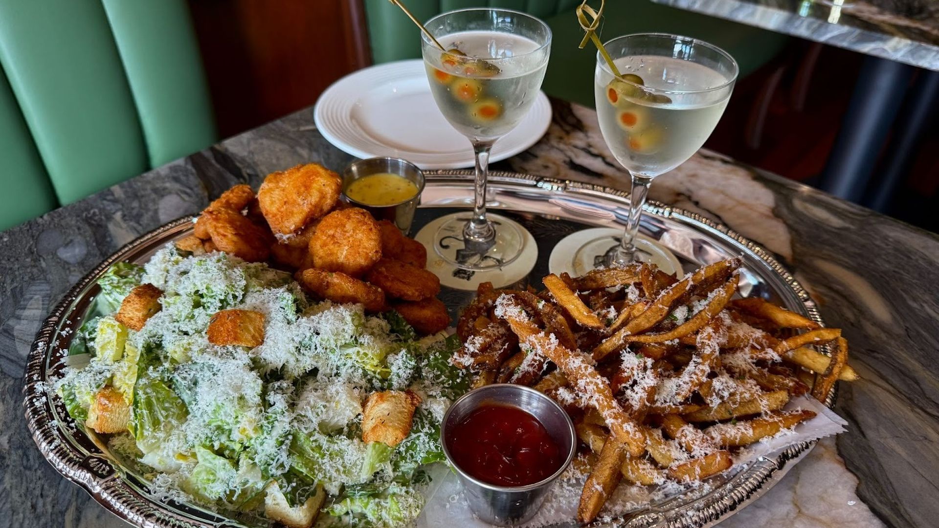 Silver platter with fried chicken nuggets, Caesar salad topped with grated cheese and croutons, crispy fries with grated cheese, dipping sauces, and two martinis garnished with olives on a marble table.
