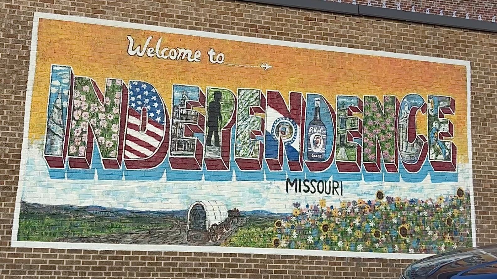 Colorful mural on a brick wall reads Welcome to INDEPENDENCE MISSOURI. The word INDEPENDENCE is a mosaic of patterns—flag stripes, flowers, and textures—over a prairie with sunflowers.