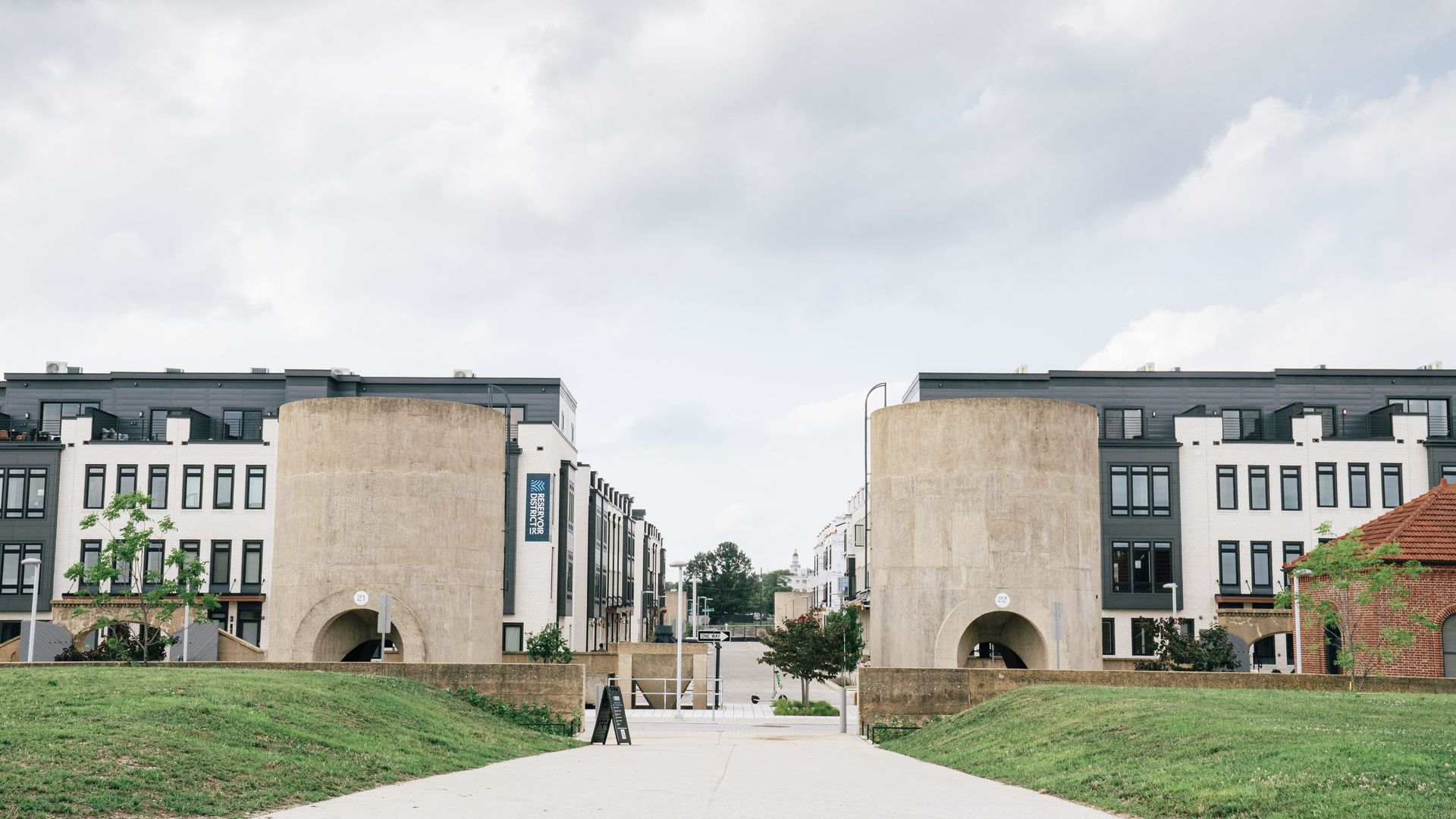 Pathway between two large beige cylindrical water filtration structures, leading to modern black and white buildings under a cloudy sky with green grass on either side.