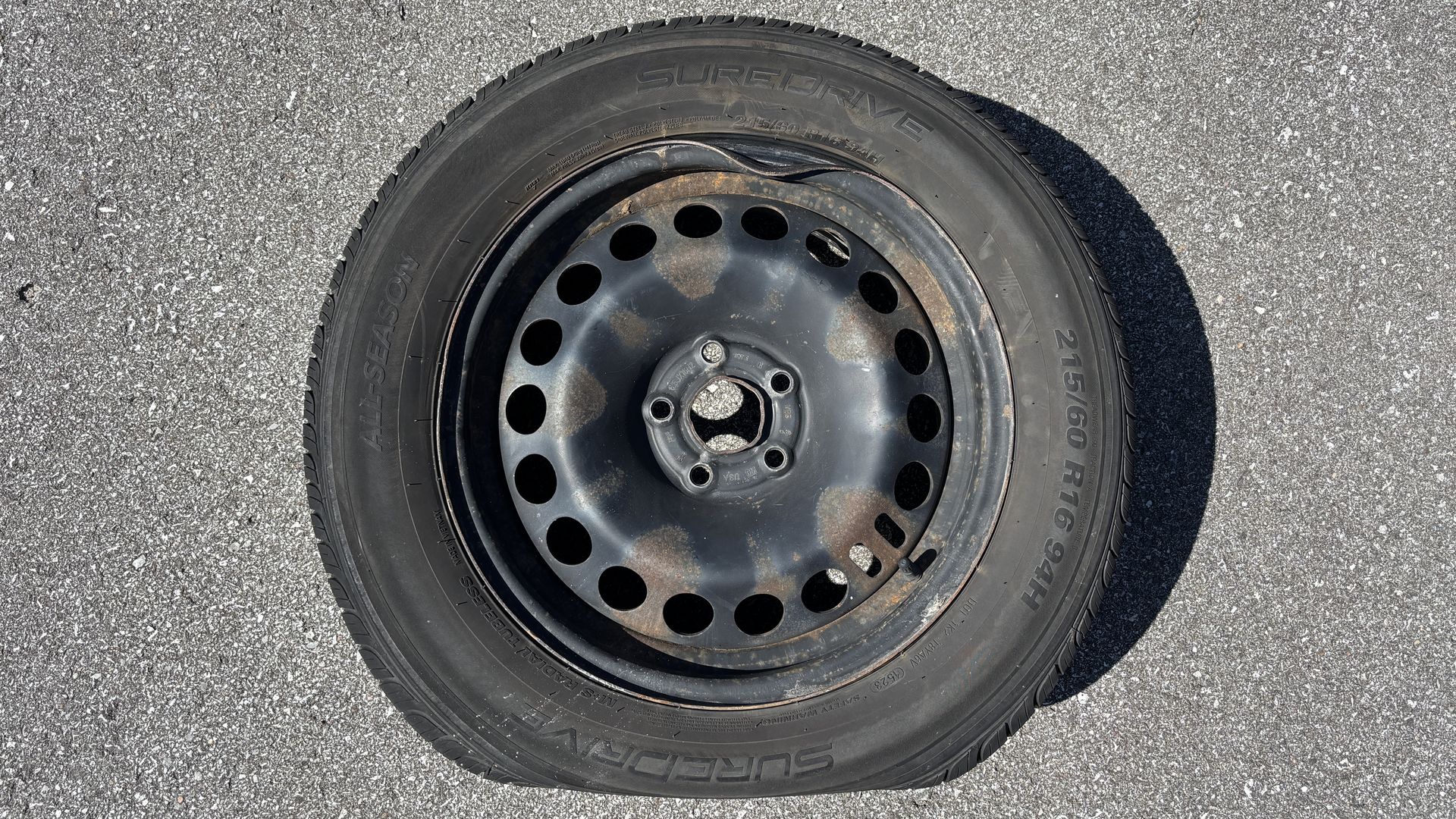 Black steel wheel with circular holes and rust spots, mounted with a tire on rough asphalt. 
