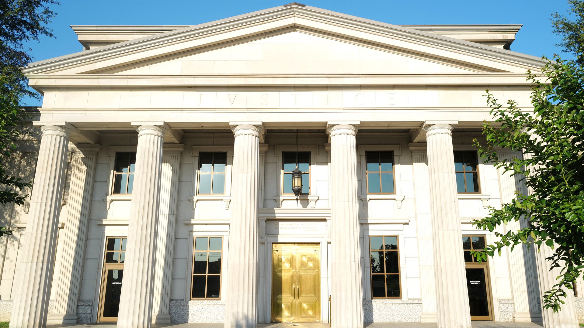 Sunlit neoclassical courthouse facade with six tall stone columns, a golden central door, and "JUSTICE" inscribed above, framed by trees under a clear blue sky.