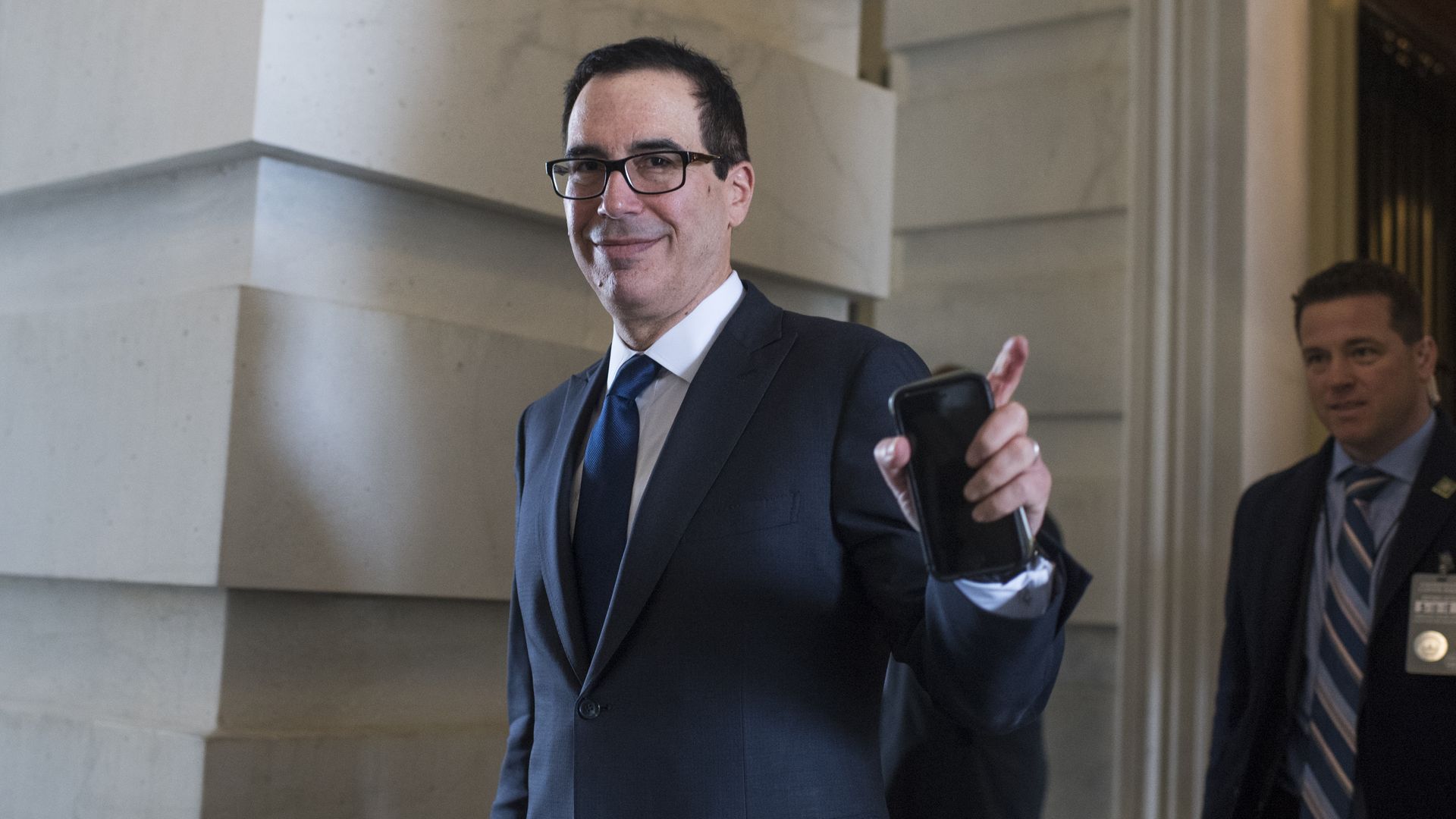 Steve Mnuchin gestures in the direction of the camera with his left hand, holding his phone, as he walks by a stone feature of the Capitol building