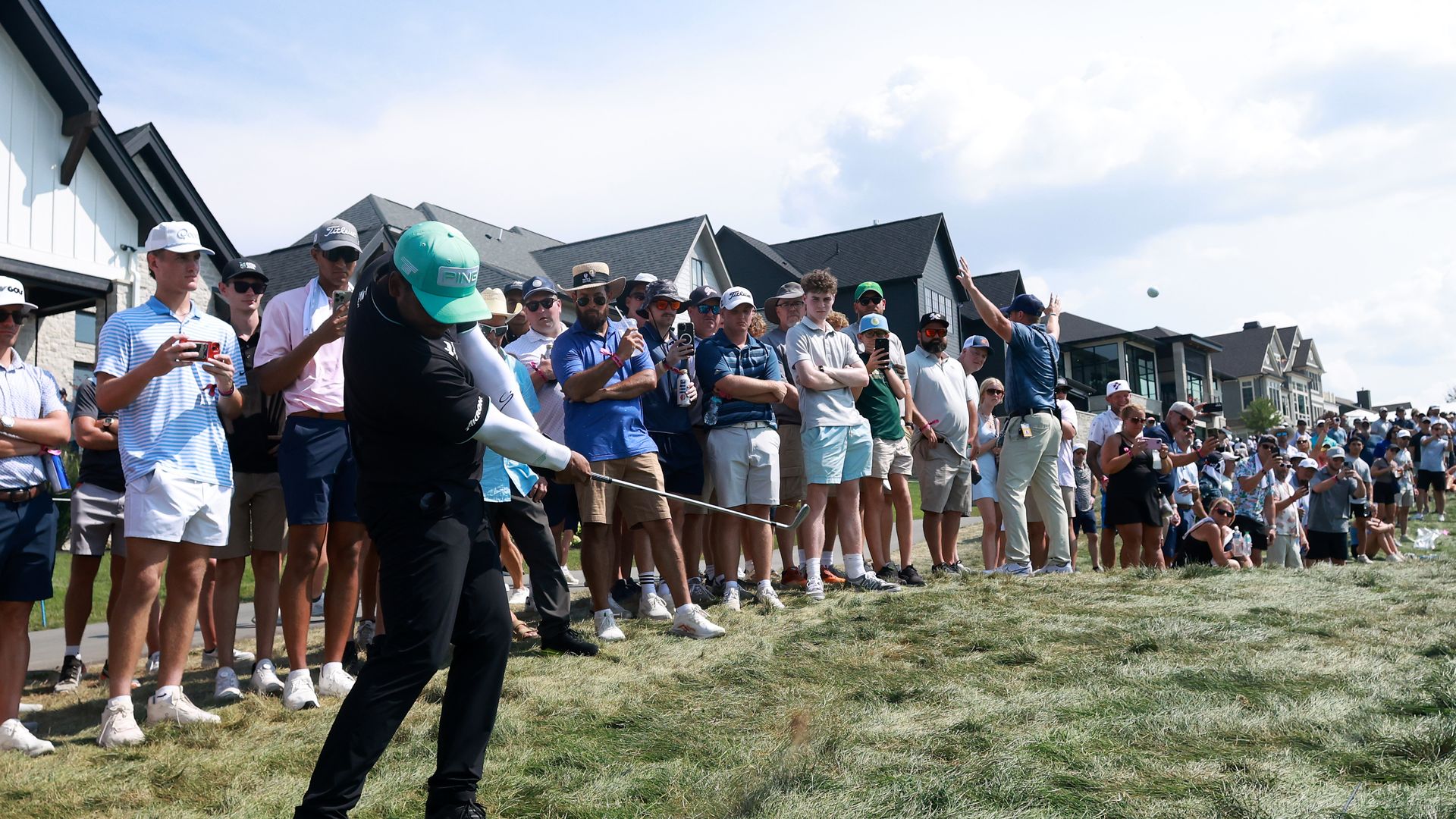 Golfer in black and green swings club on grassy area in front of large crowd of spectators near houses under blue sky with clouds.