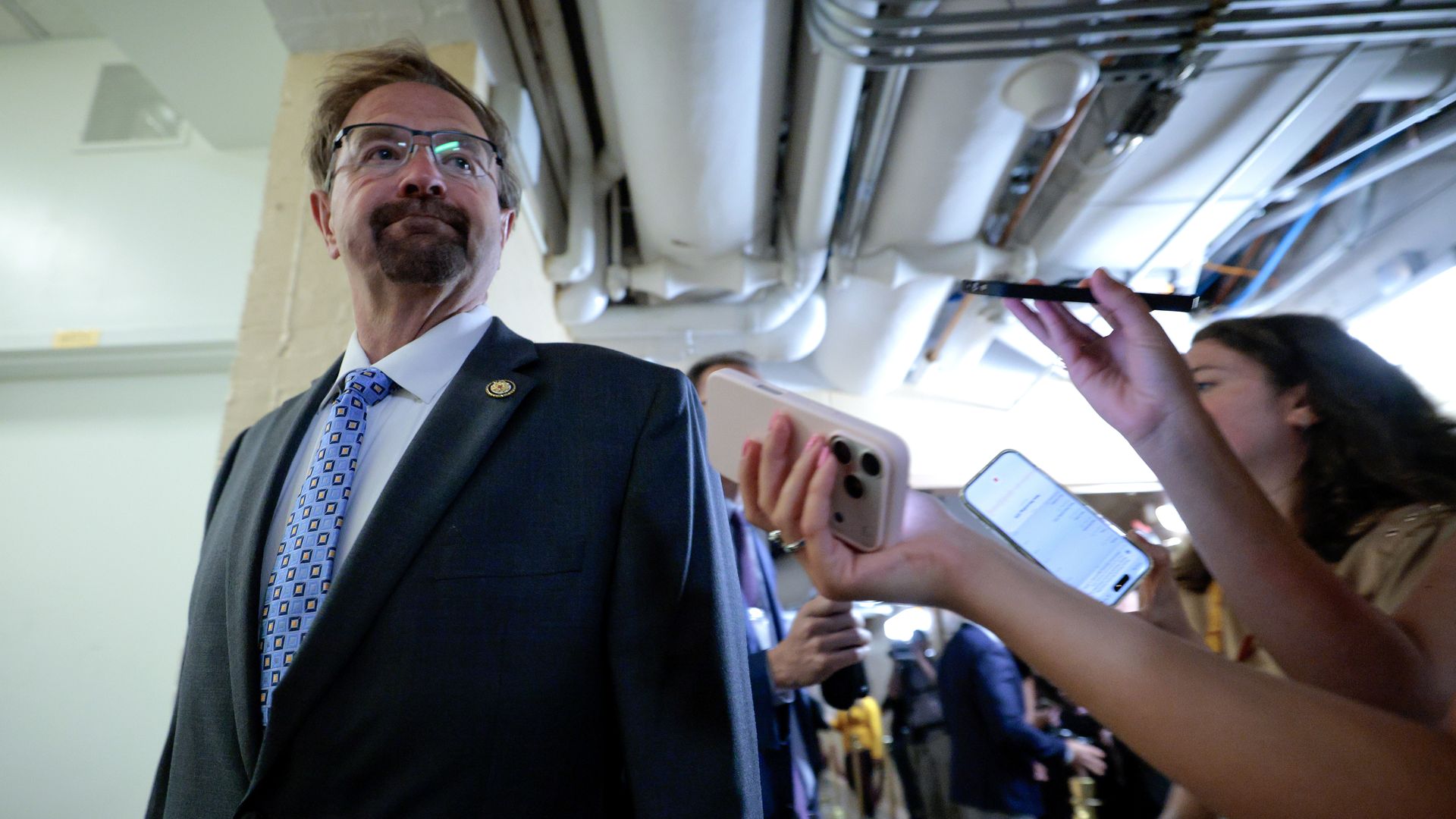 Man in a dark suit with a blue patterned tie and glasses stands in a hallway with exposed pipes as reporters press smartphones and a microphone toward him.