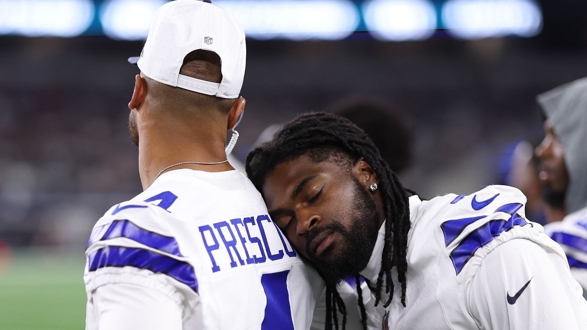 One Dallas Cowboys player with a white cap and jersey labeled PRESCOTT leans while another player with black dreadlocks rests his head on Prescott’s shoulder during a game.