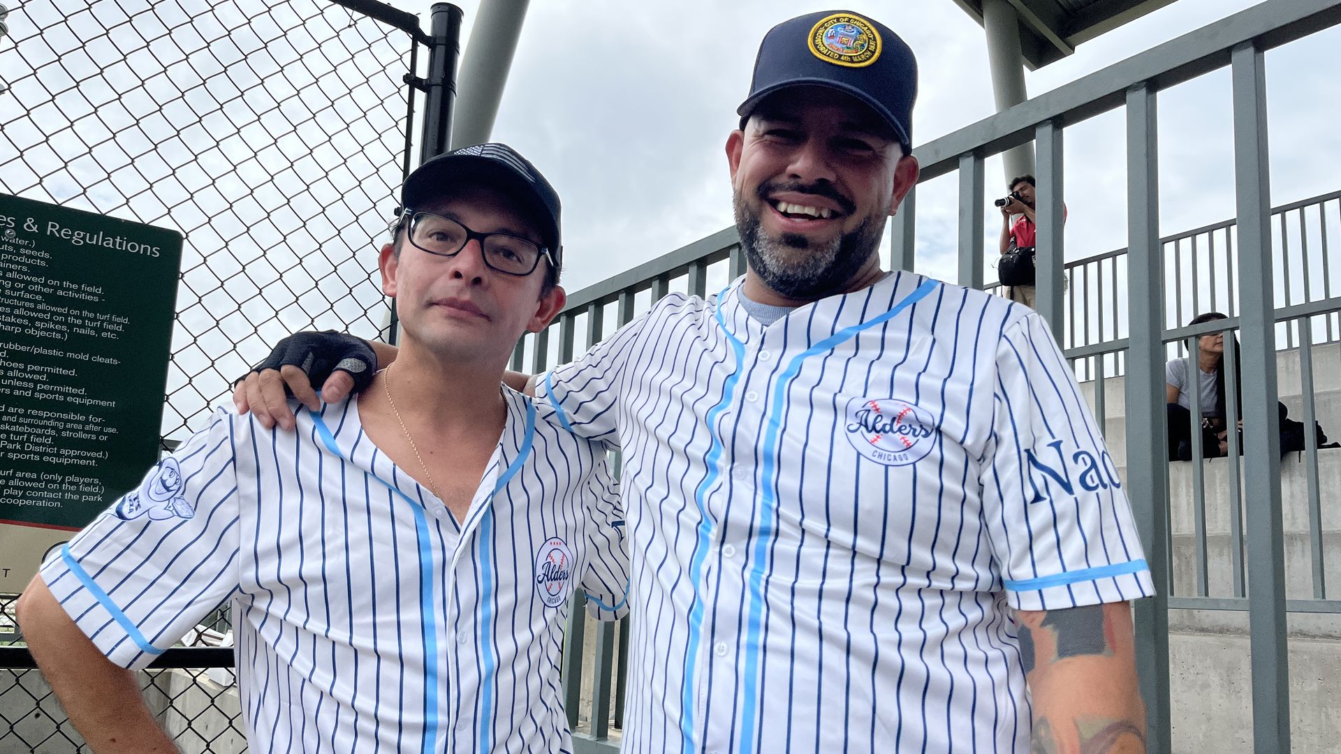 Two men wearing white baseball jerseys with blue pinstripes and black caps stand by a metal fence, one with glasses and the other smiling, with a tattoo on his arm.