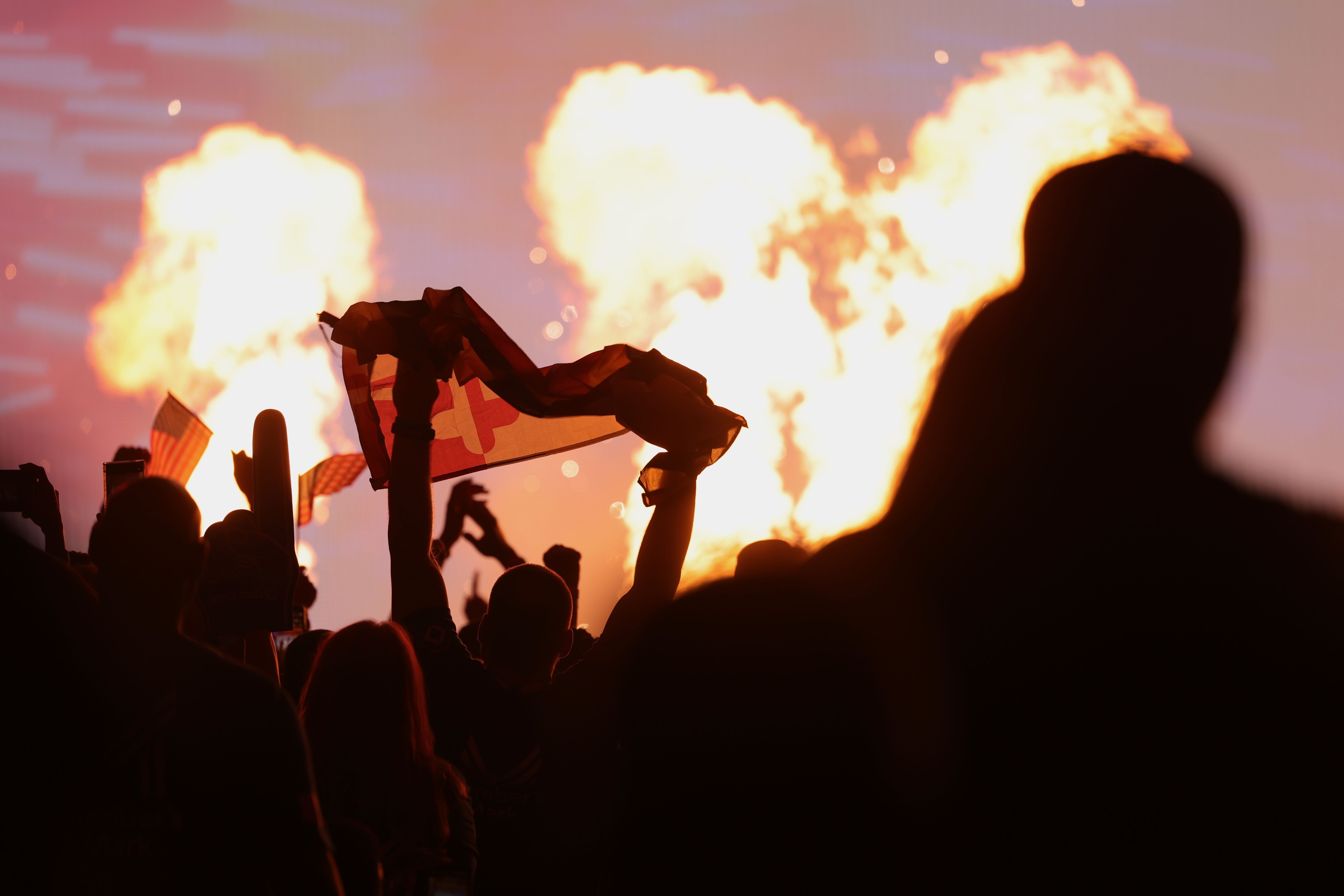A fire explodes on a stage with people cheering in the foreground.