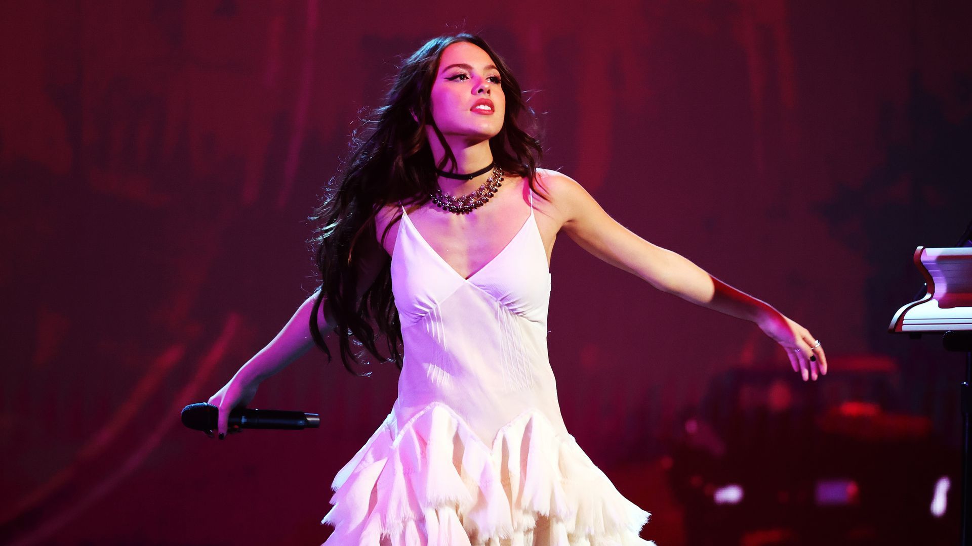 Olivia Rodrigo on stage in a white dress with her arms out and a microphone in her right hand
