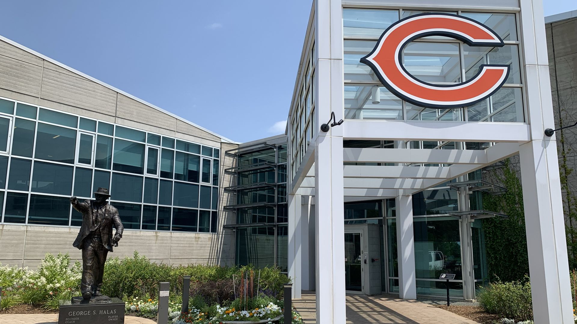 Entrance to a glass and concrete building with a large orange and white Chicago Bears logo above. A statue of George S. Halas stands to the left, surrounded by colorful flowers.