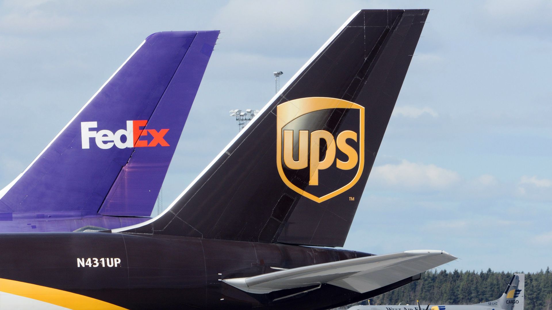 Two airplane tails on a tarmac: left purple FedEx tail, right dark brown UPS tail with a gold shield logo; airport equipment and more planes visible in the background.