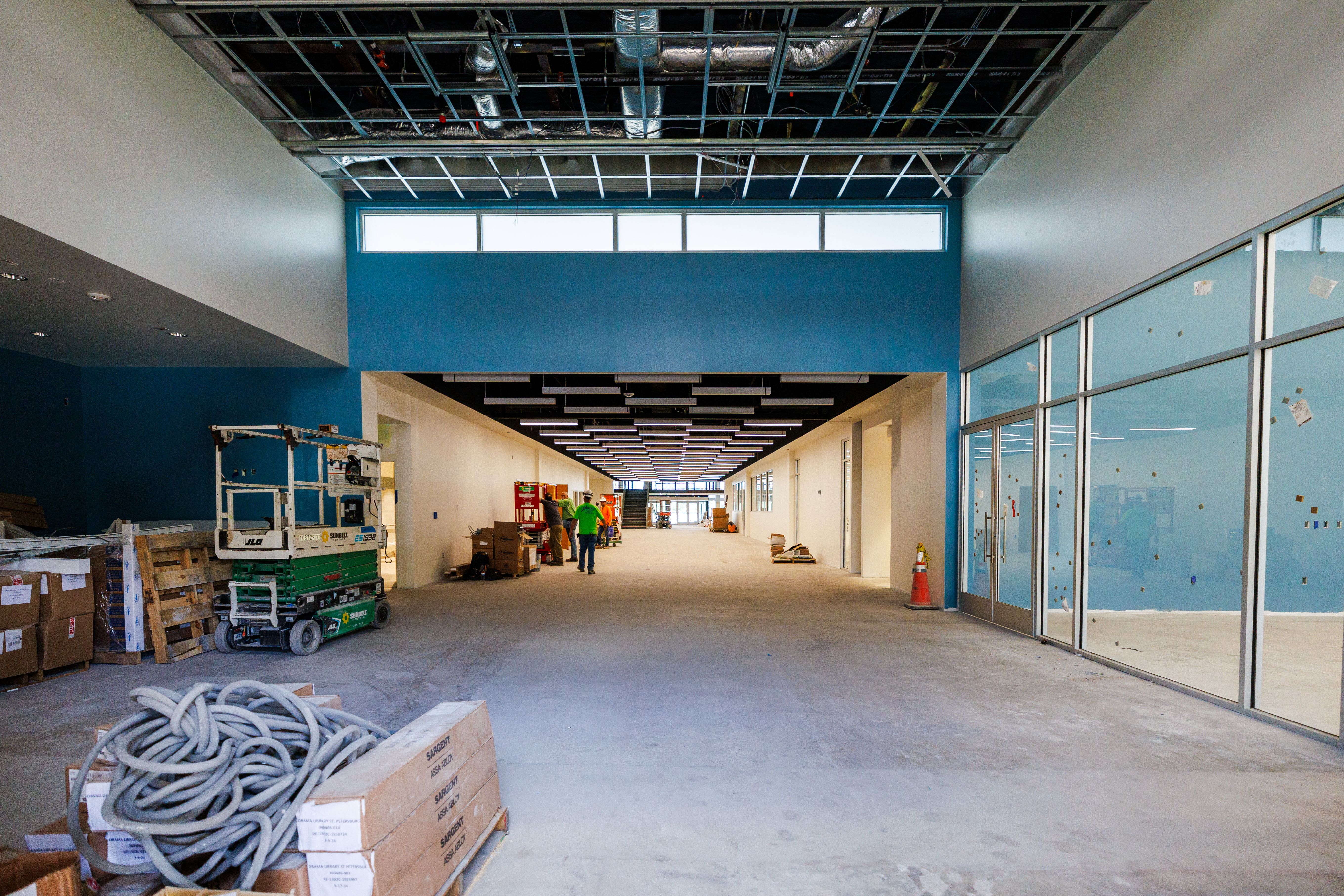 A large, wide hallway with blue and white walls. Construction equipment covered in dust is scattered around.