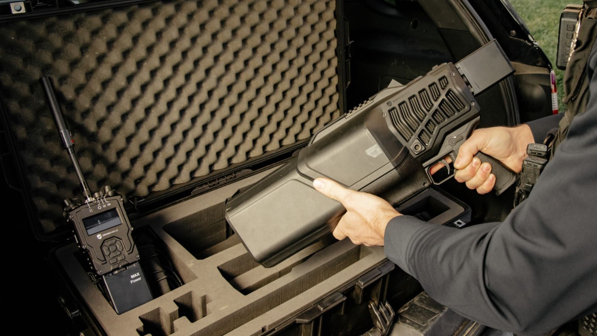 A person in dark clothing handles a large, tactical device with a grip, placing it into a foam-padded hard case; a control unit with a display sits nearby.