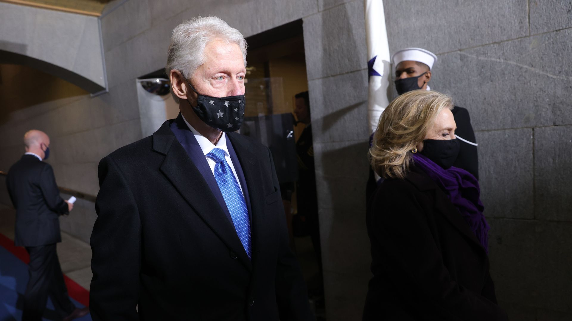 Photo of Bill Clinton wearing a mask and in a coat walking in a hall