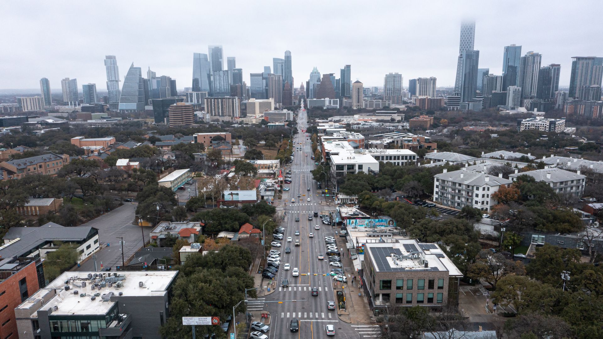 Aerial view of a downtown skyline with tall glass towers, a central avenue lined with trees and cars, and low-rise buildings in the foreground on an overcast day.
