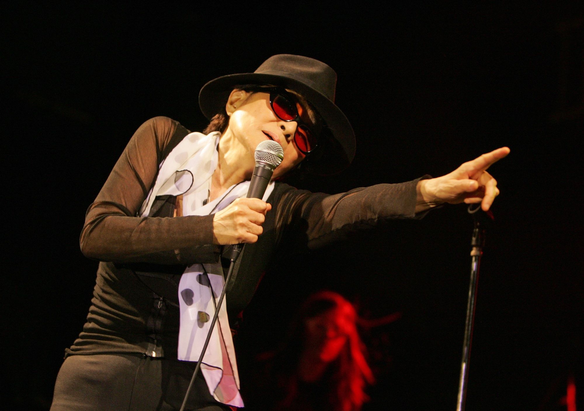 Yoko Ono in a black hat and sunglasses singing into microphone and pointing off stage.