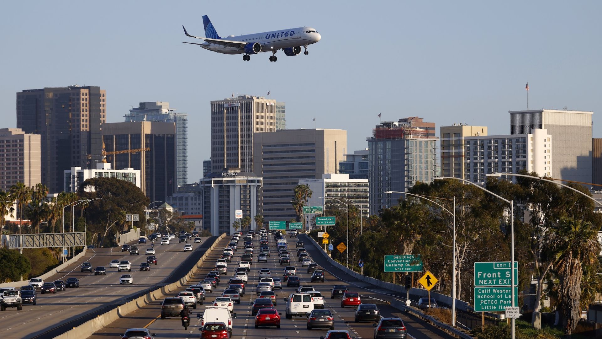 A United Airlines plane flies over buildings and the freeway with traffic heading into downtown San Diego. 