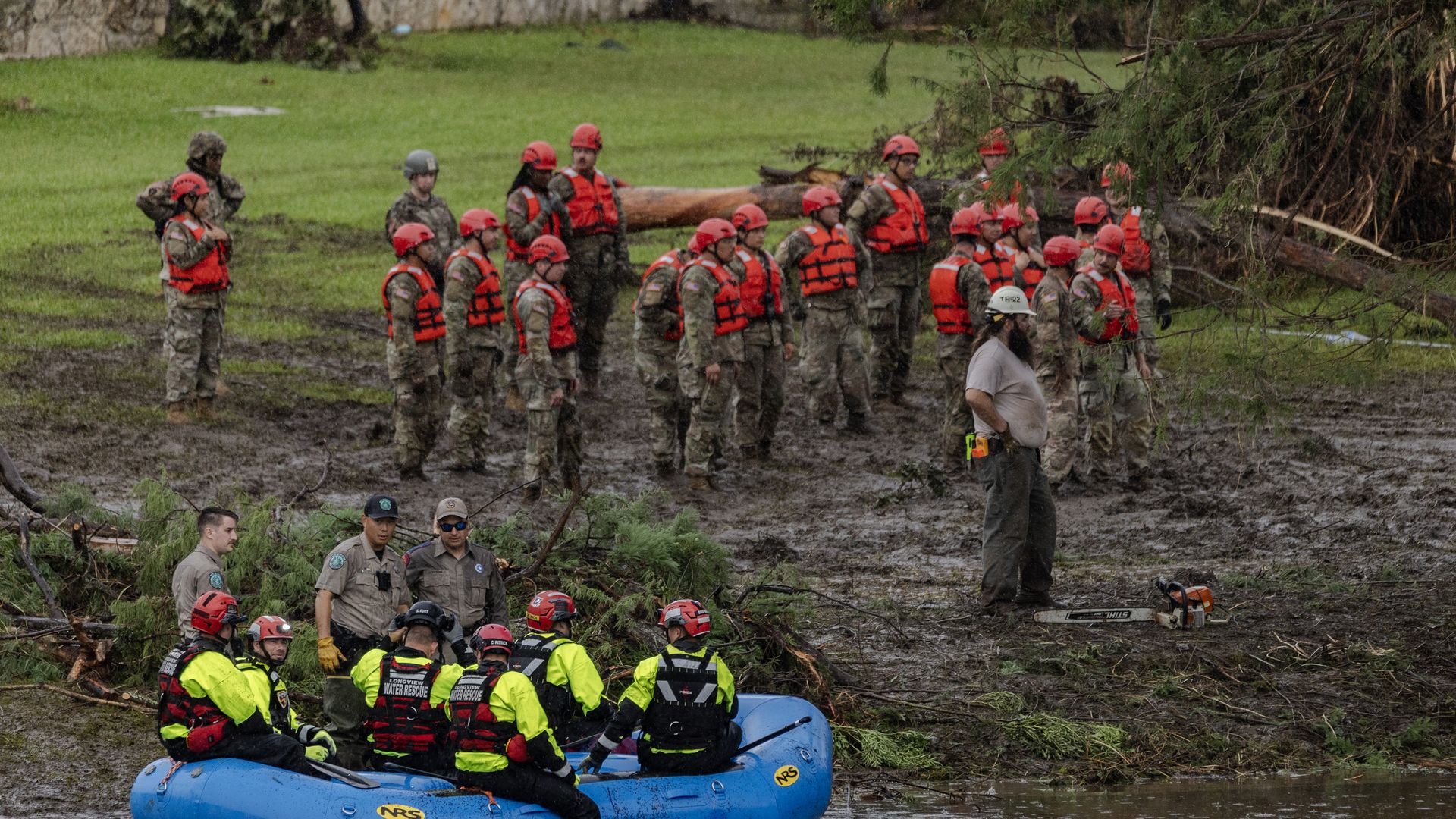 several emergency workers in camouflage and helmets and vests stand near the remnants of the Central Texas floods. A blue boat carrying several rescuers is next to them
