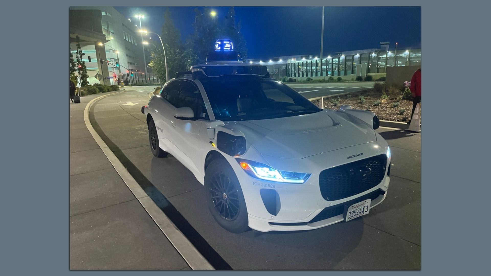 White Waymo robotaxi with sensors, and a blue lidar unit on the rooftop, parked at a hotel curb at nighttime, with building lights in the distance. 