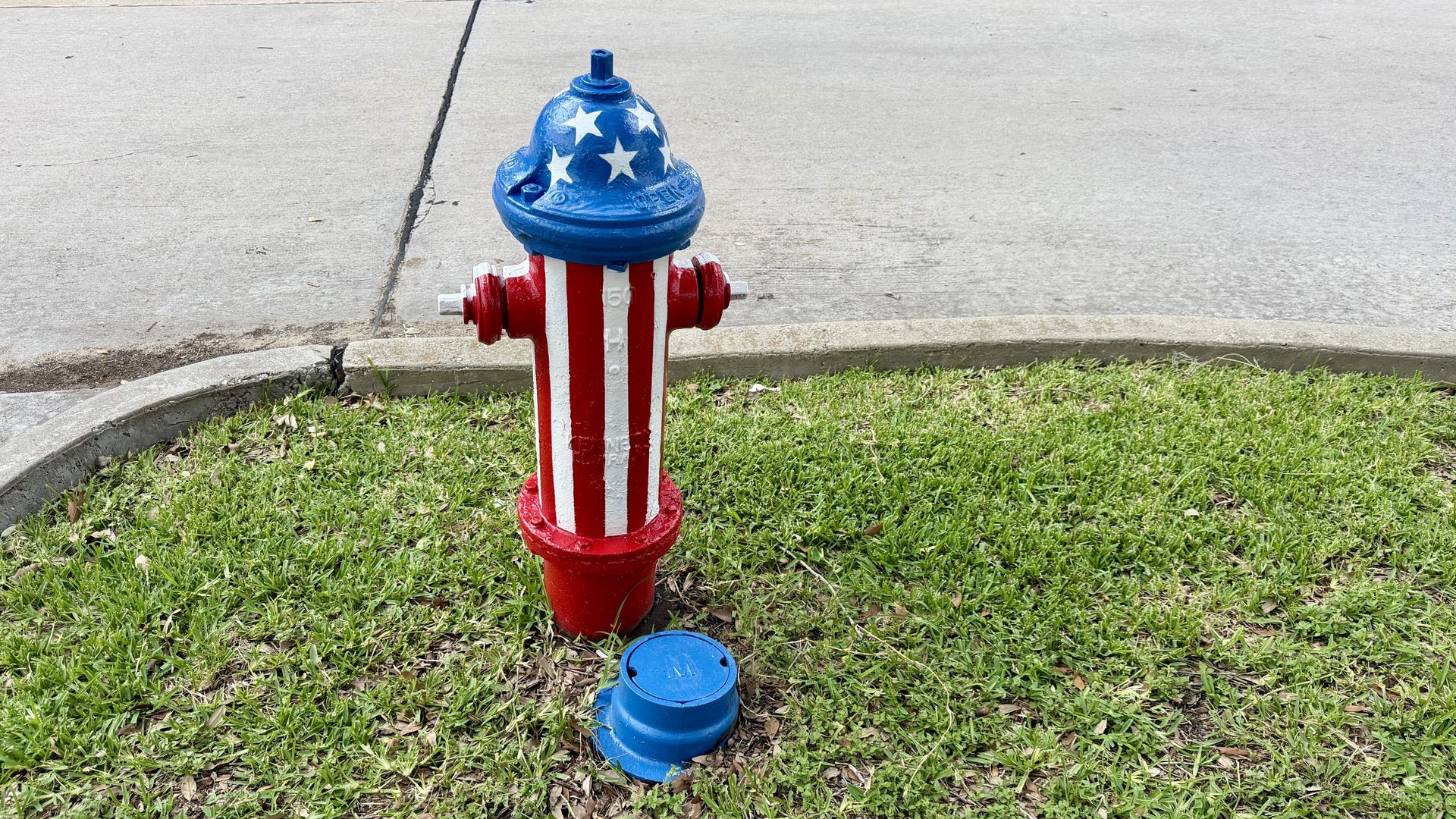 Fire hydrant painted like the American flag - blue top with white stars, red and white striped body - standing on a grassy verge beside a sidewalk.