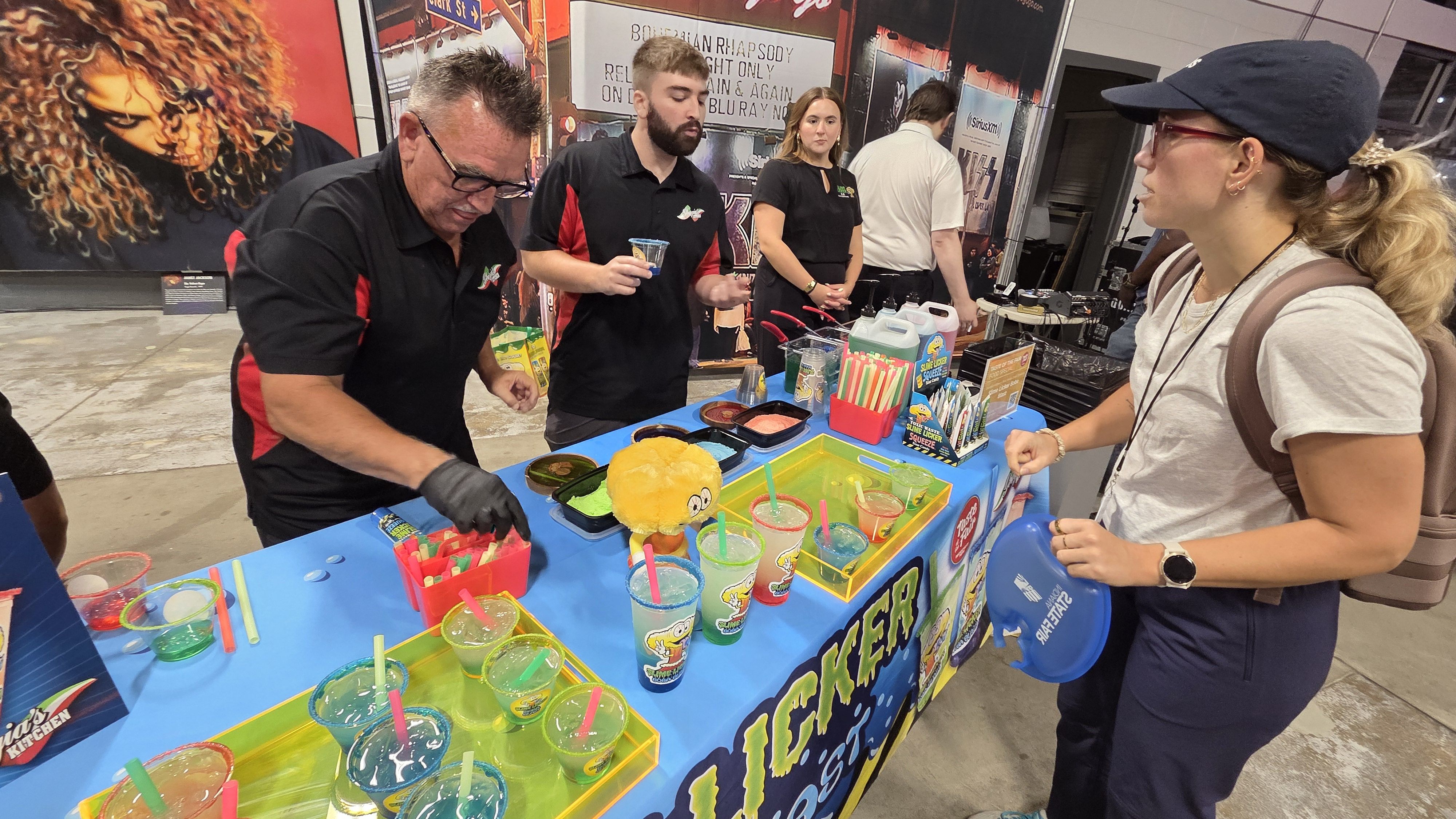 People at a colorful booth serving vibrant drinks with straws and green, orange, and blue slimes in containers. One person wears black gloves, another holds a cup, and a woman with a cap and backpack stands nearby.