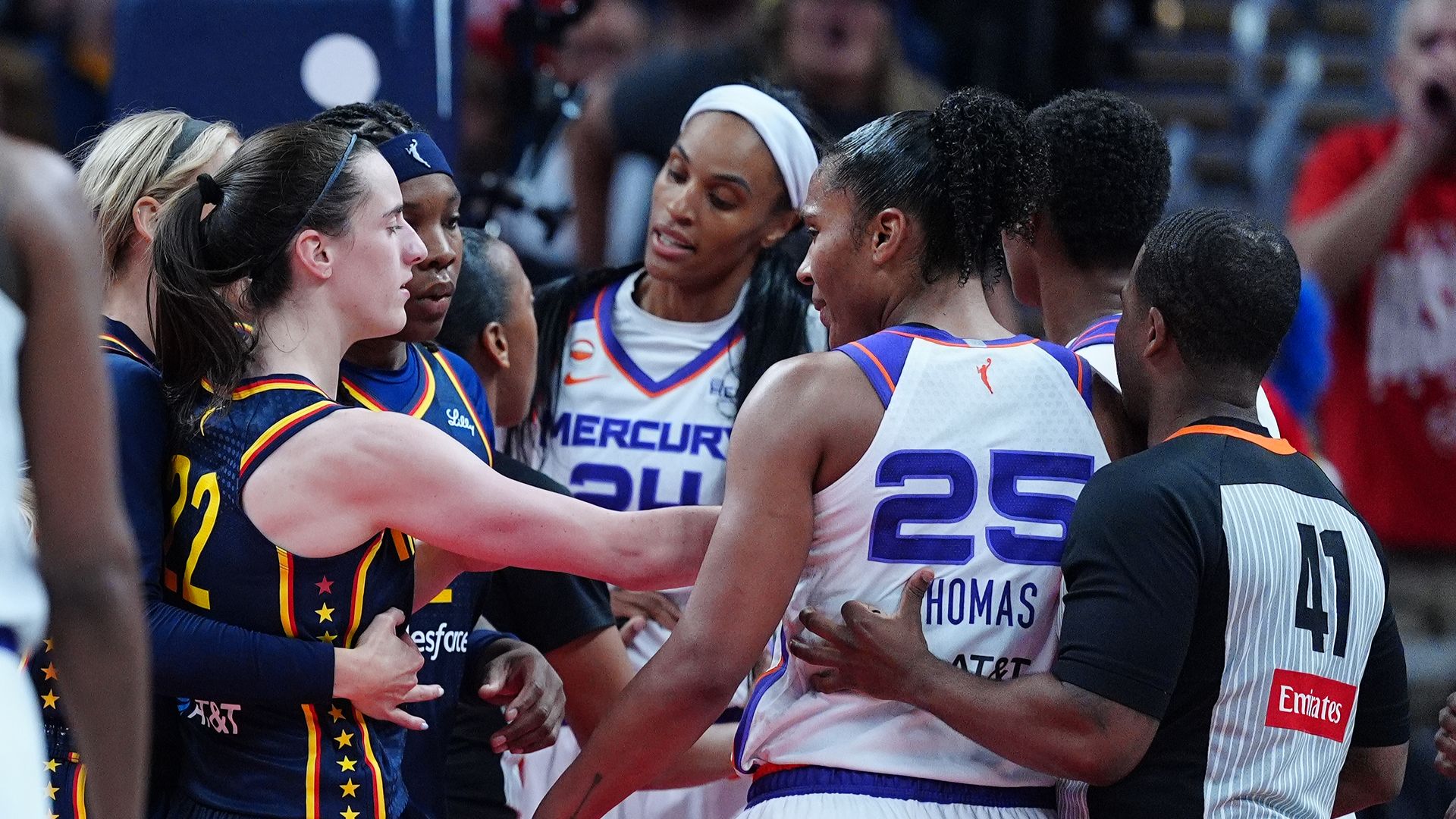 A group of female basketball players in a huddle on the court, arms around each other. Center player wears a white headband and white-and-purple jersey; others wear blue uniforms with stars.